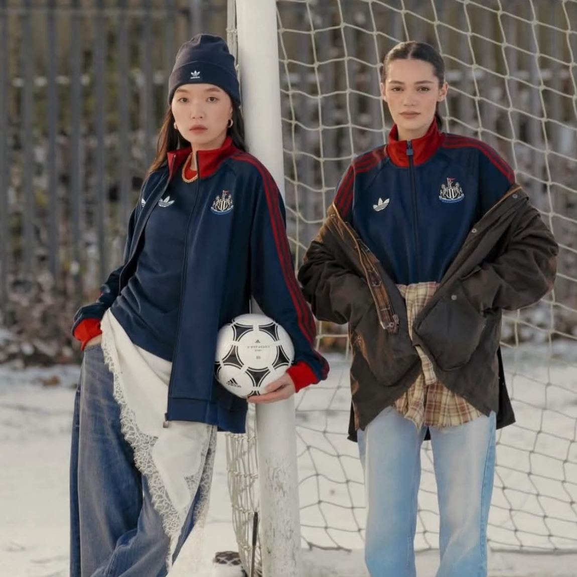 Two young women stand by a soccer goal on a snowy field, dressed in athletic jackets, with one holding a soccer ball.