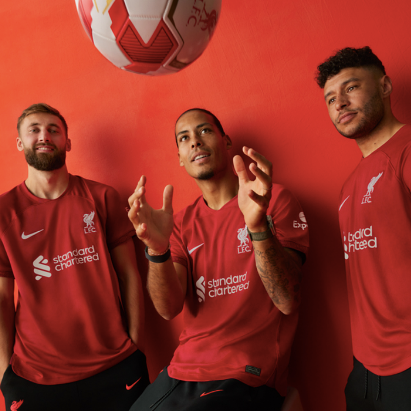 Three men in red Liverpool Football Club jerseys standing against a red wall, one of whom is tossing a soccer ball in the air.