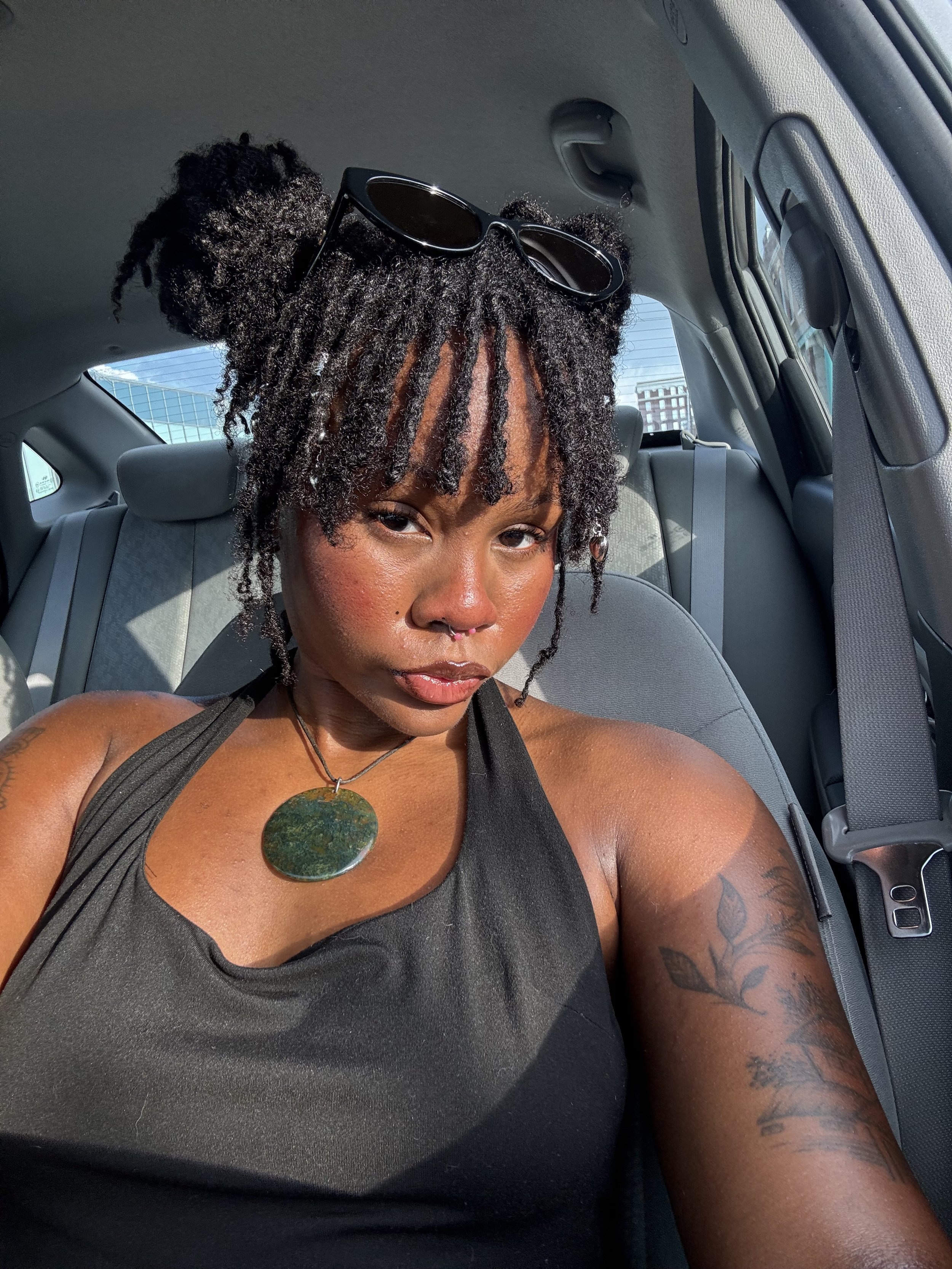A woman with dreadlocks sitting in the backseat of a car, wearing sunglasses on her head, a tattoo on her arm, and a necklace with a large circular pendant.