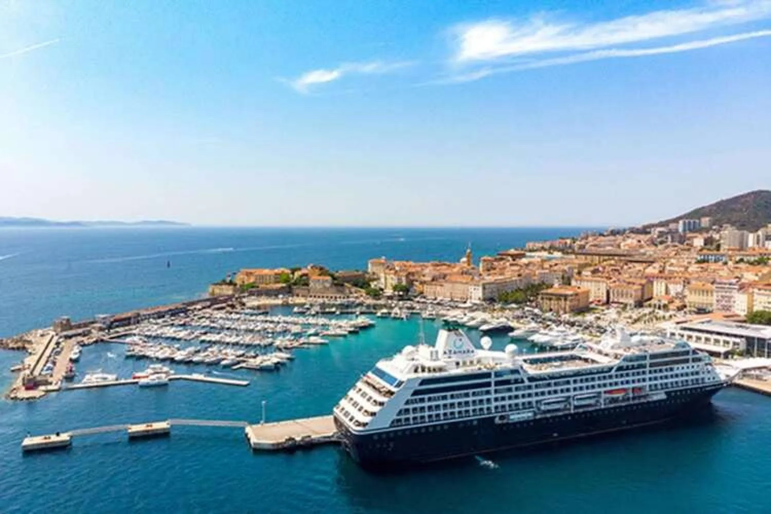 Aerial view of a marina with a cruise ship docked, surrounded by boats and a coastal city with buildings and hills under a blue sky.