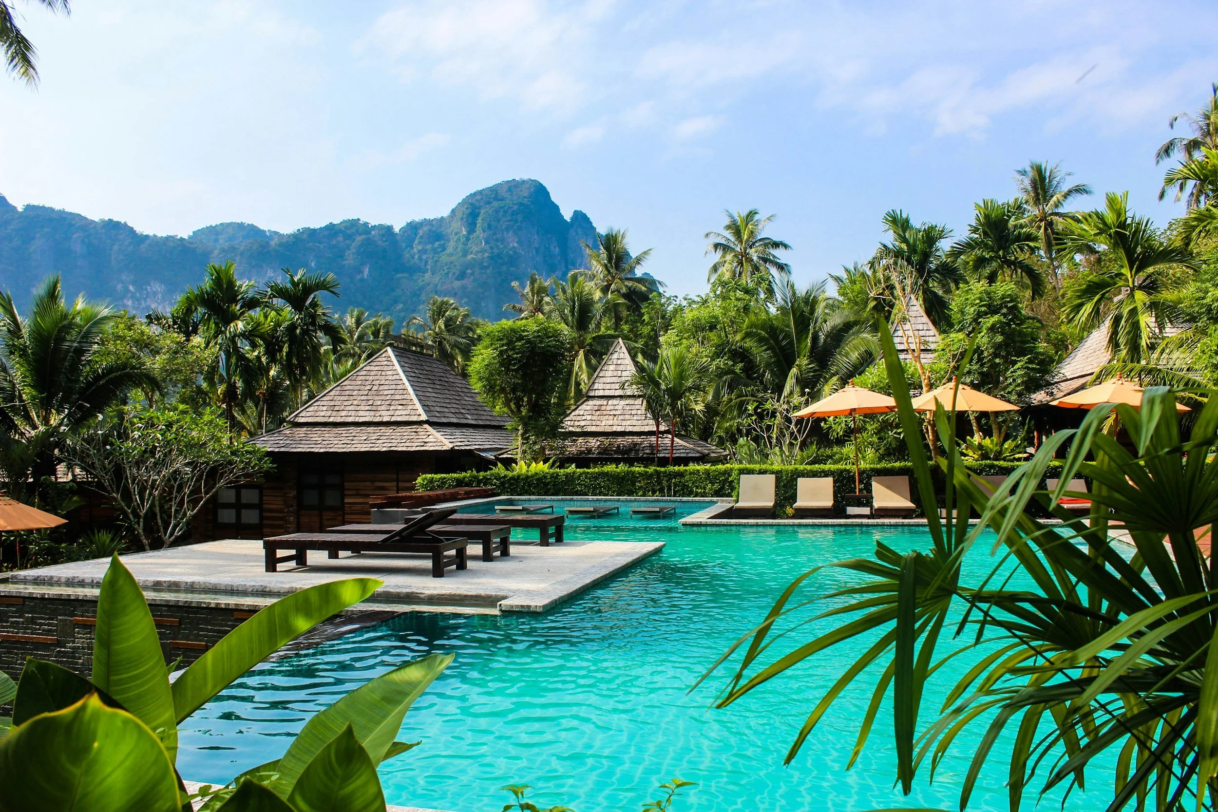 Swimming pool area with lounge chairs, surrounded by tropical plants and huts, with mountains in the background under a blue sky.