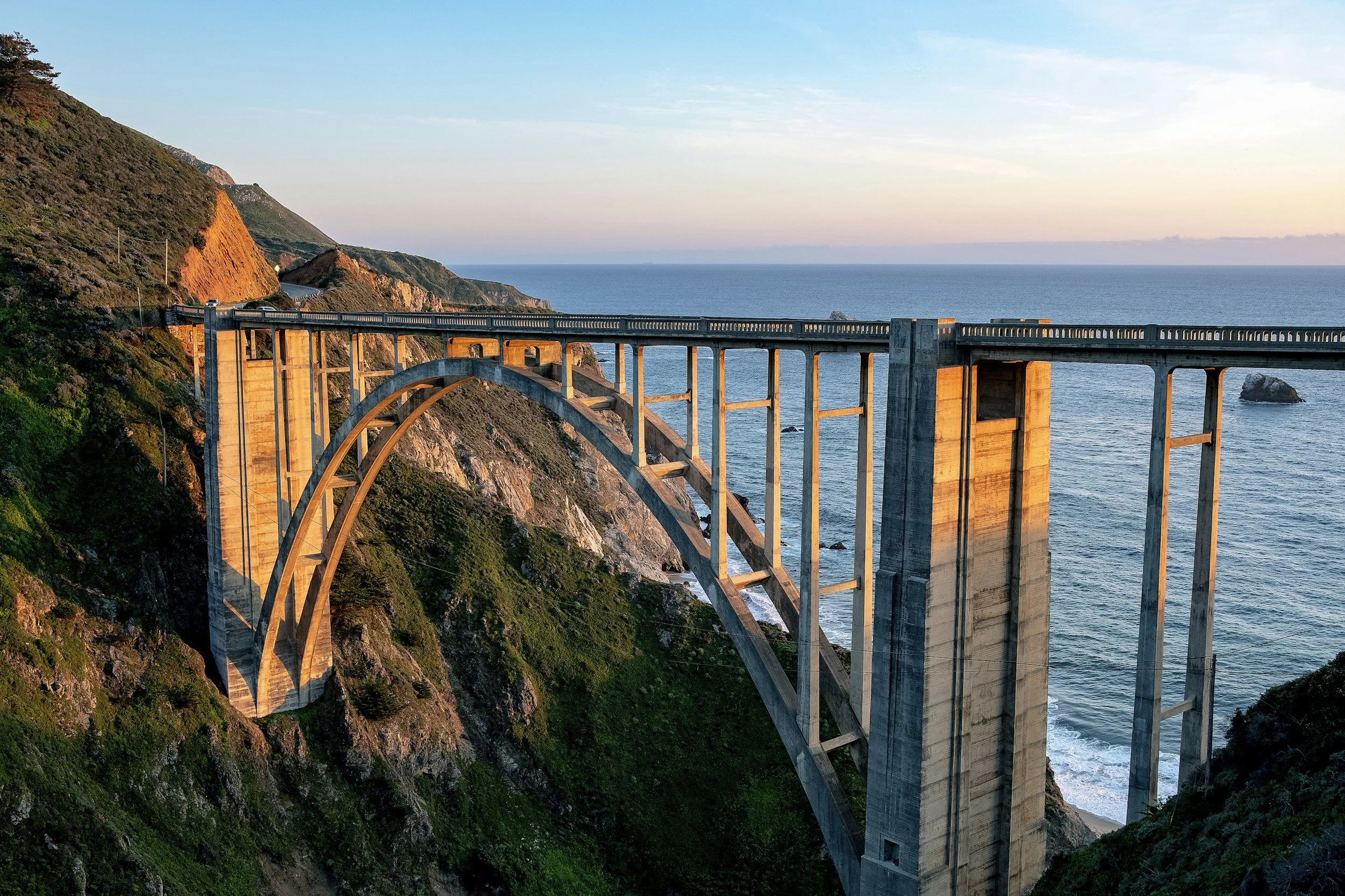 Bridge over a cliffside along the ocean, with coastal hills and a sunset sky in the background.