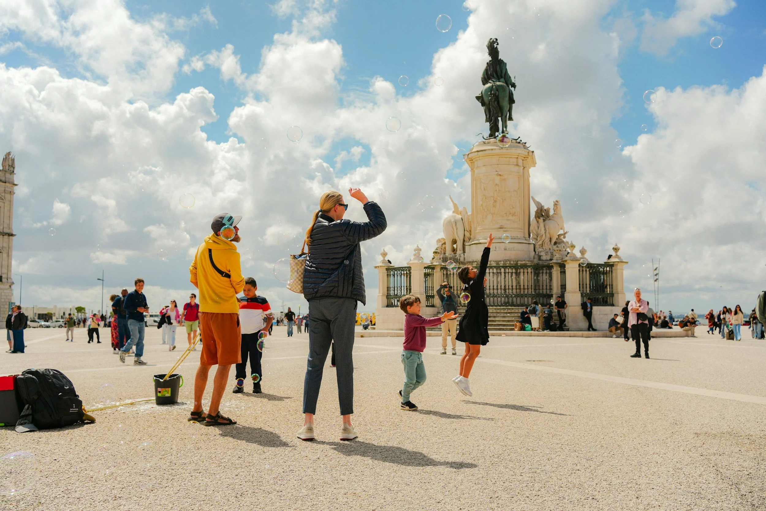 People playing with bubbles in a plaza with a large monument of a man on horseback in the background, under a partly cloudy sky.