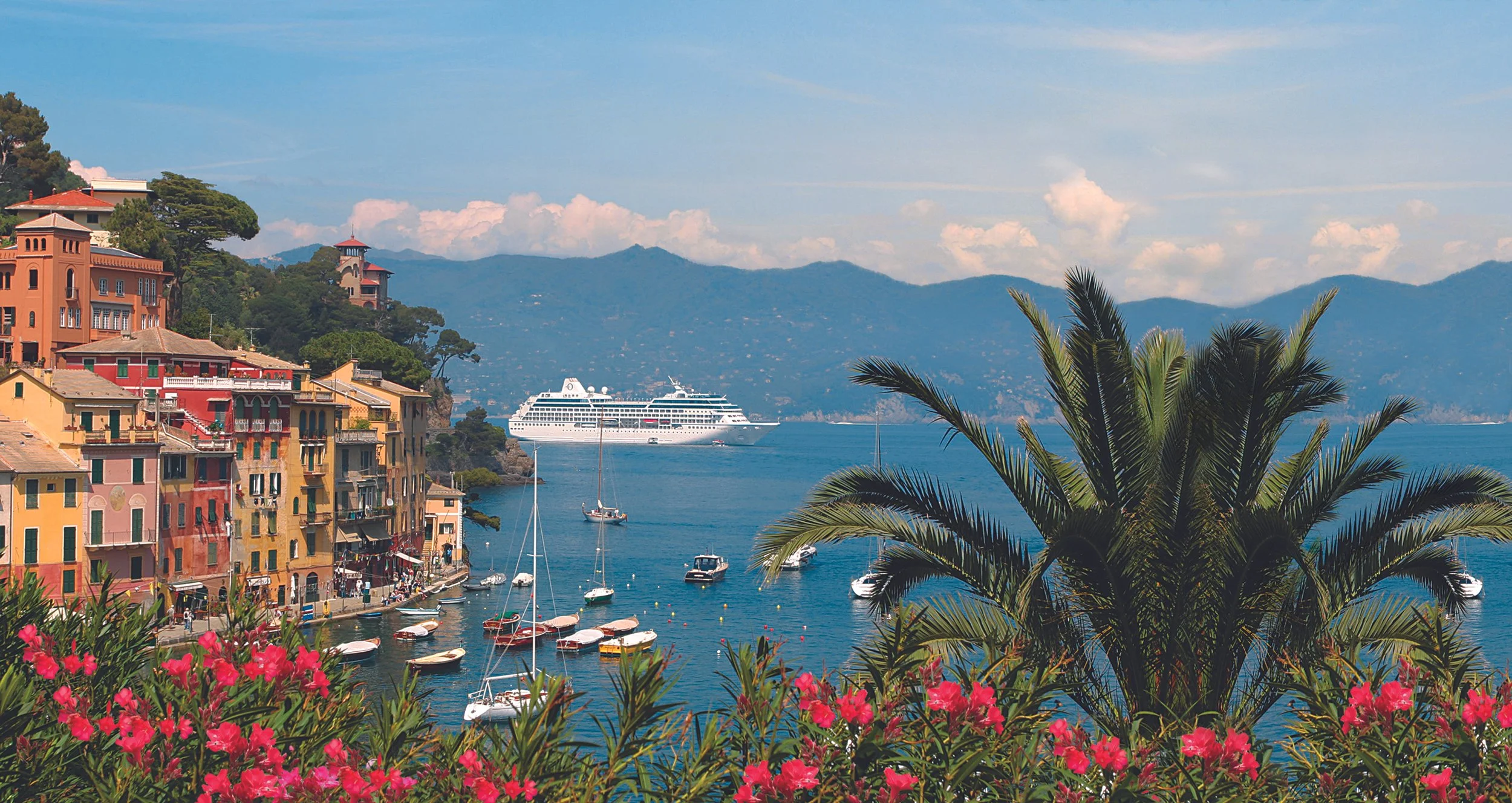 Colorful buildings along a lakeside with boats anchored in the water, a cruise ship in the background, mountains and a partly cloudy sky, and palm trees and pink flowers in the foreground.