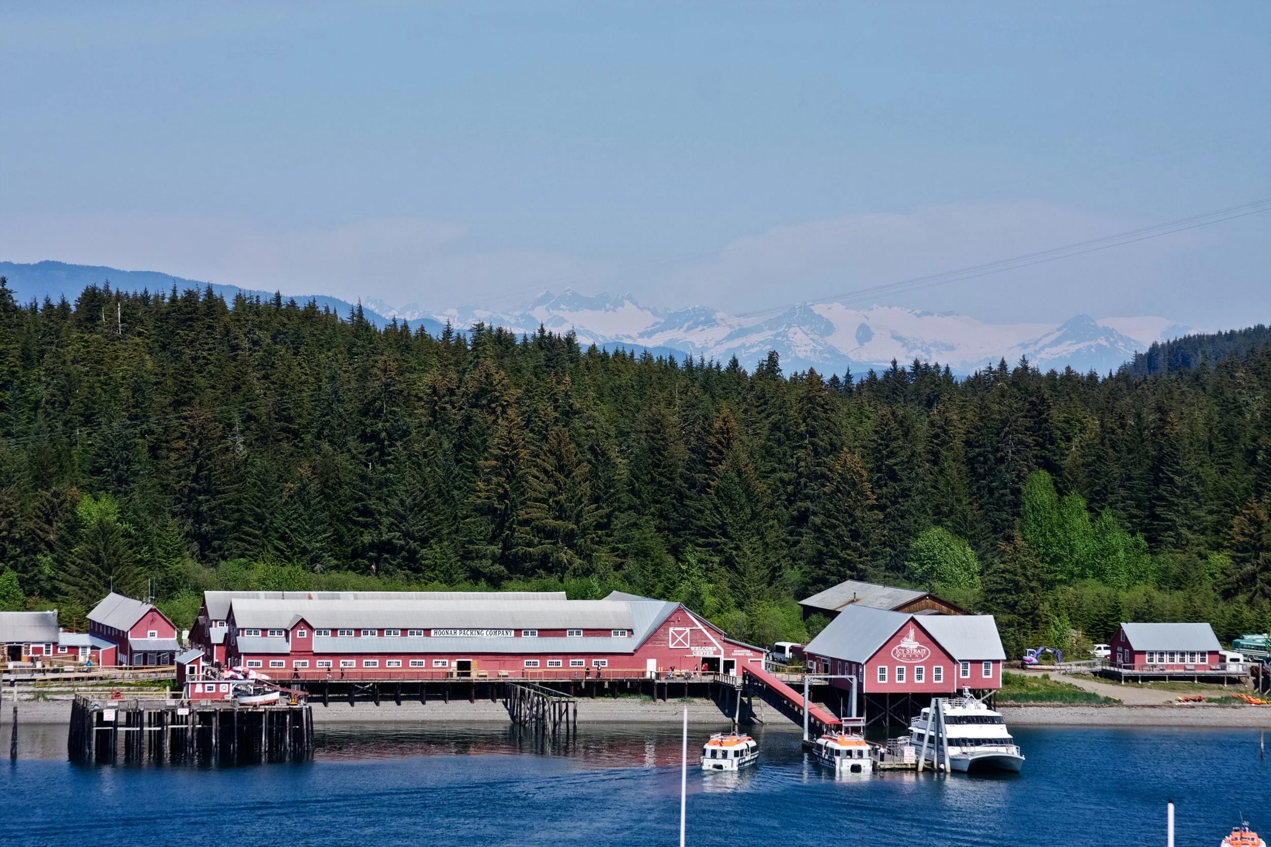 A waterfront scene with a red and white dockside building labeled 'Hoodah Packing Company' on a pier, boats docked in the water, lush green trees covering hills, and snow-capped mountains in the background under a light blue sky.