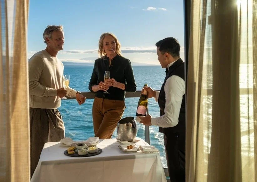 Three people on a cruise ship deck enjoying champagne, with the ocean and sky in the background, as a waiter pours champagne into a glass.