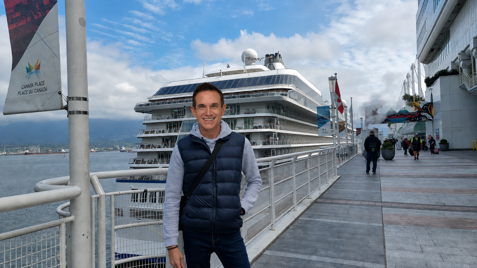 A man in a blue vest and hoodie stands on a dock in front of a large cruise ship, with other people walking nearby and the city skyline in the background.