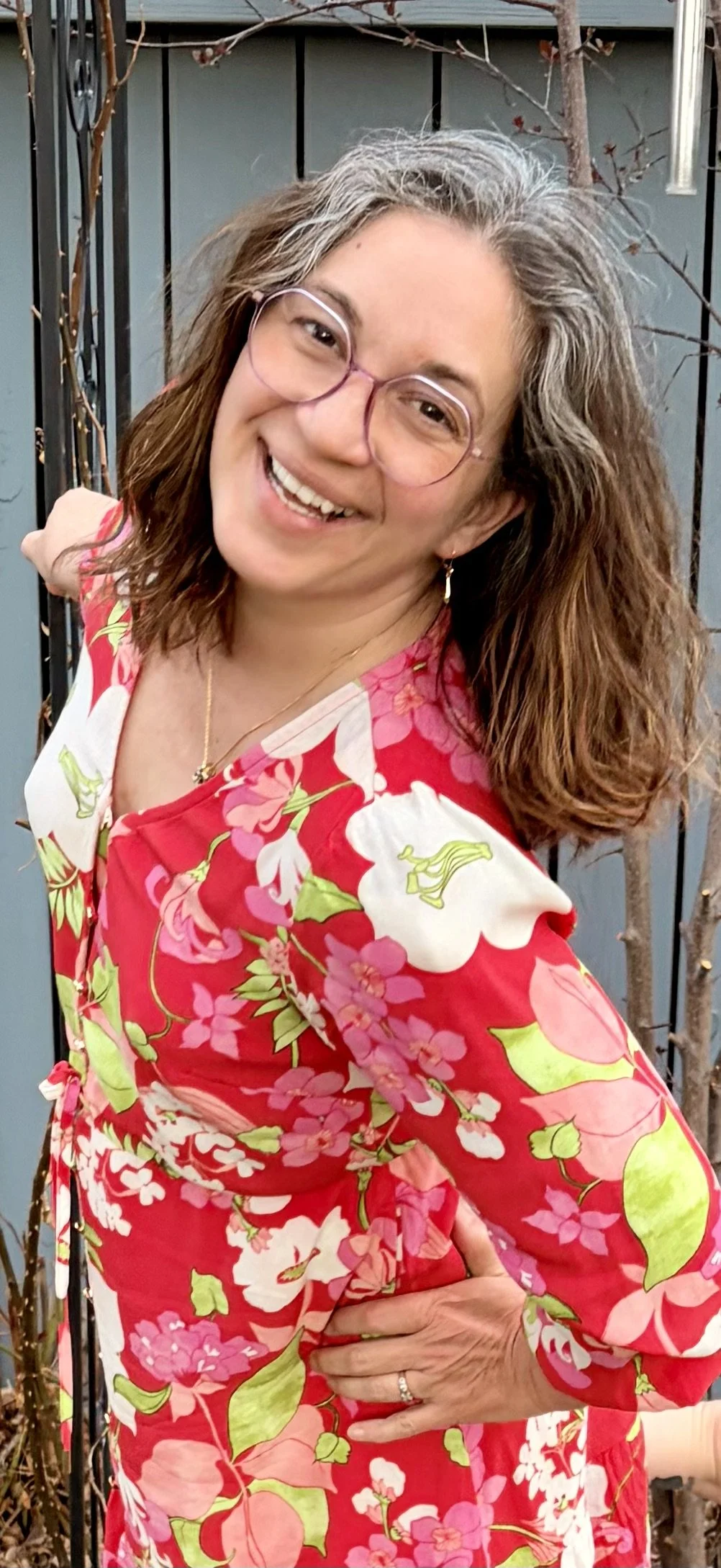 A woman with long, curly brown and gray hair smiling, wearing glasses, earrings, a ring, a necklace, and a bright red floral dress with pink, white, and green flowers, standing outdoors near a black metal fence and some leafless branches.