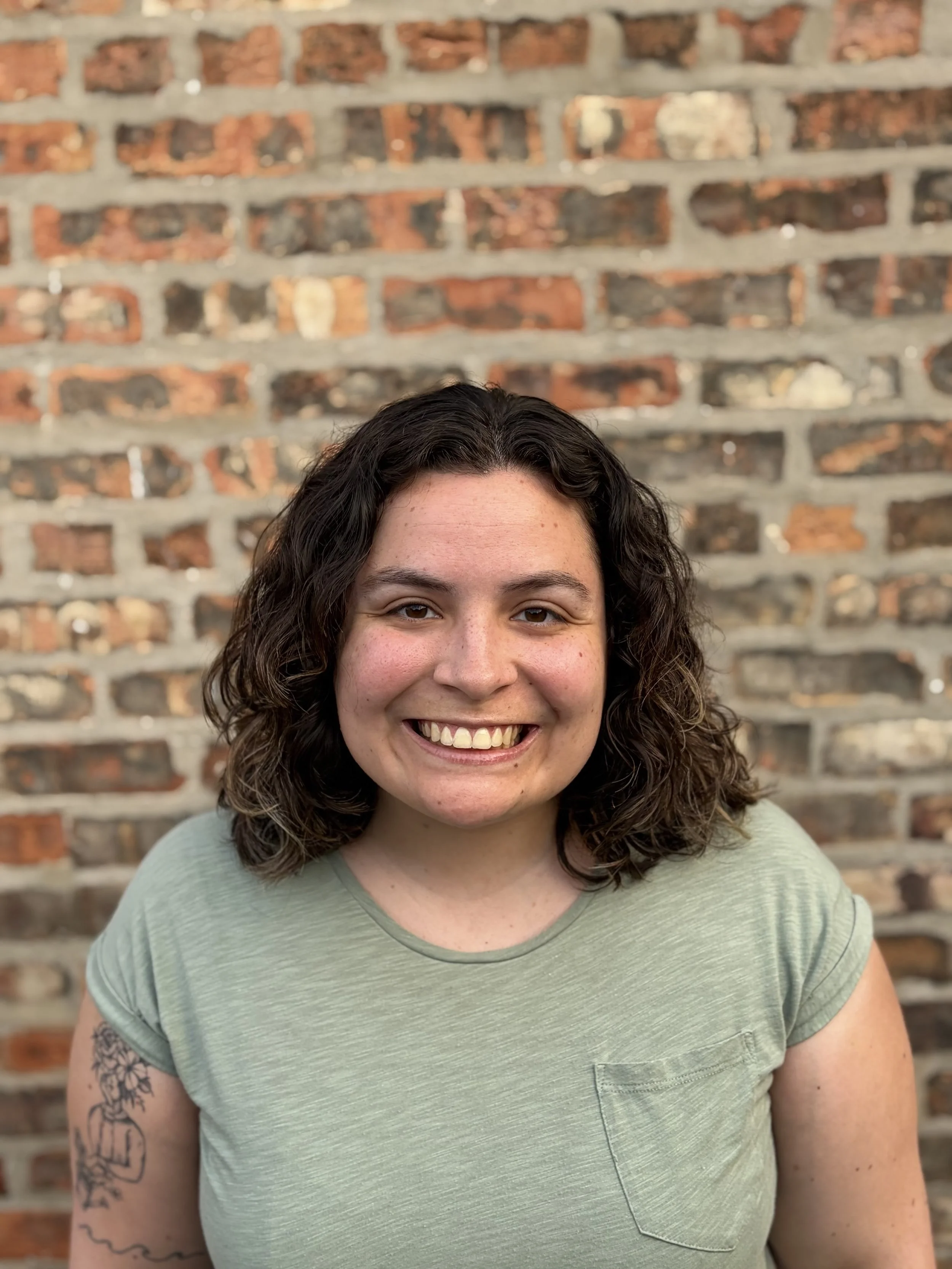 Close-up of a woman with curly brown hair, smiling, standing in front of a brick wall.