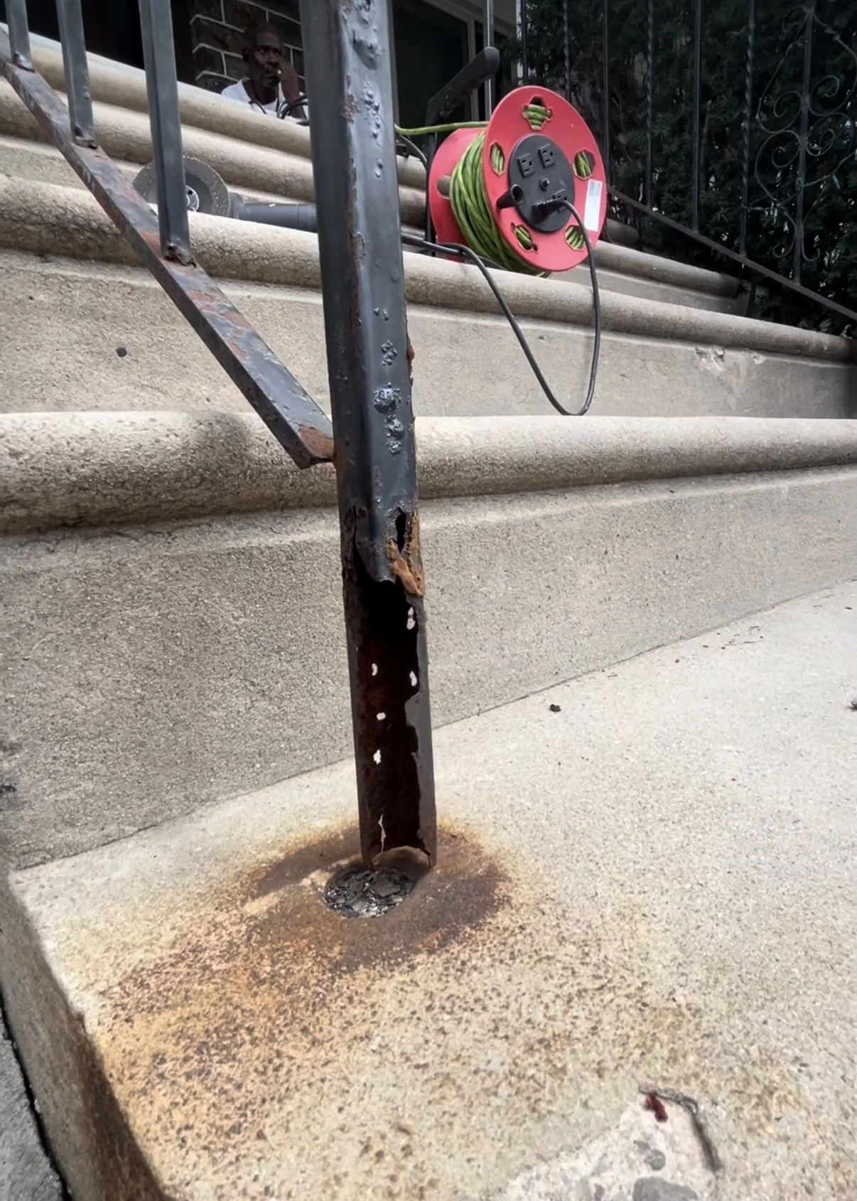 A rusty metal handrail post on concrete stairs with a power extension cord reel resting on the stairs nearby. There is a person partially visible in the background, standing behind the stairs.