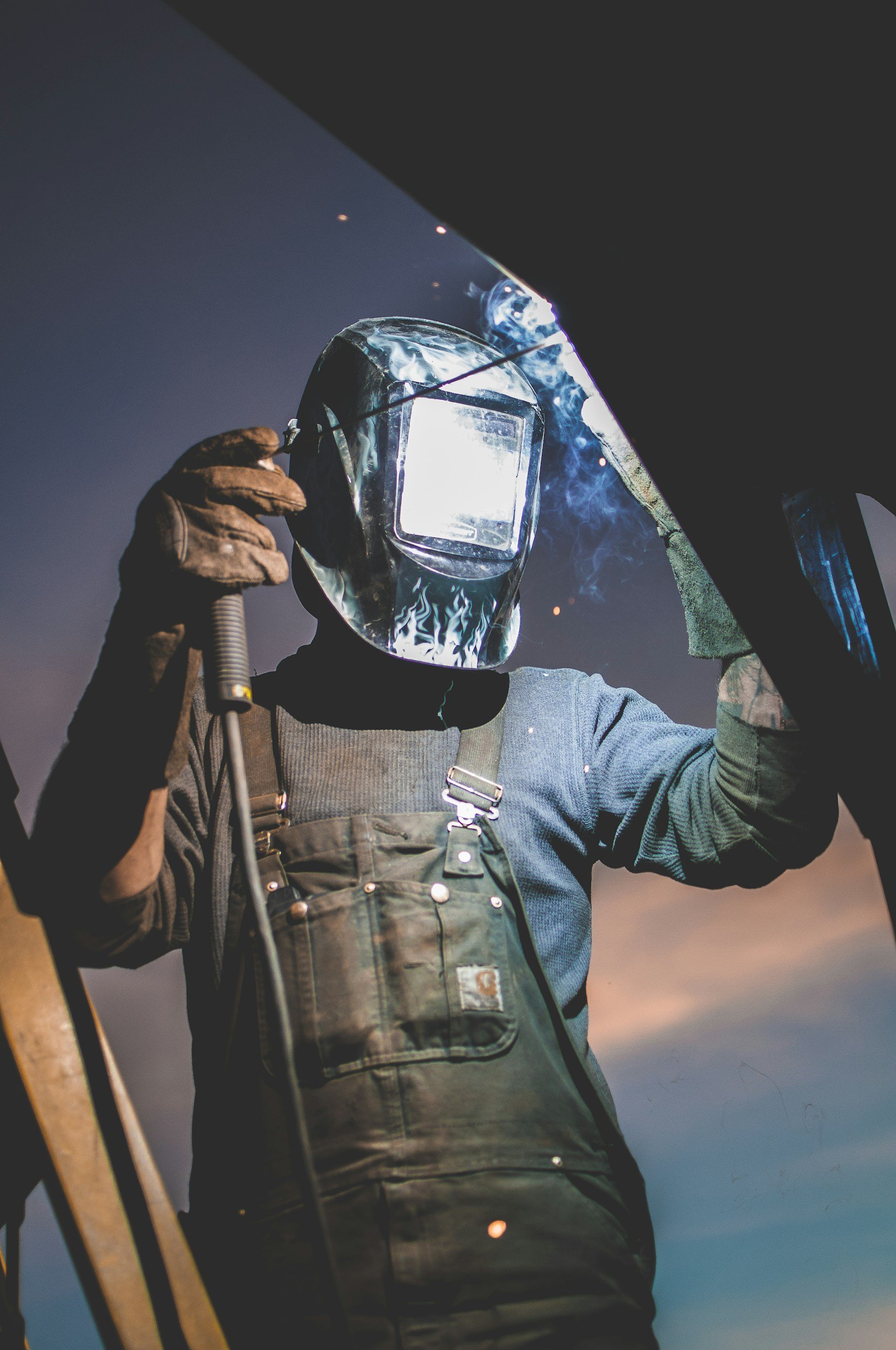 A person wearing protective welding gear, including a face shield and gloves, is welding metal outdoors at dusk or dawn.