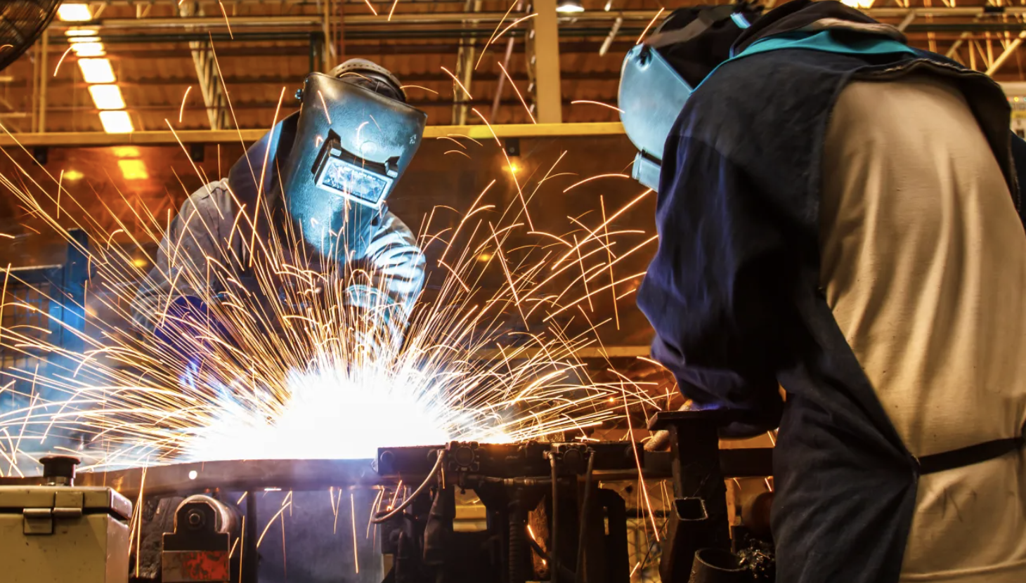 Two workers welding metal, with bright sparks flying in a factory or industrial setting, both wearing protective helmets and clothing.