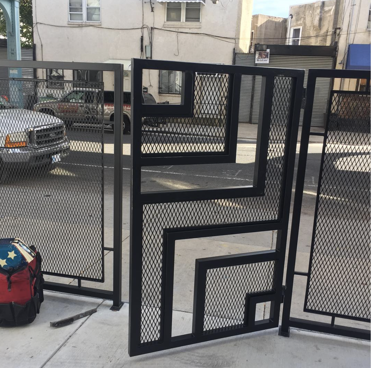 Black metal decorative folding screen with geometric patterns, set up outdoors on a sidewalk, with a red and blue bag and a hammer on the ground nearby.