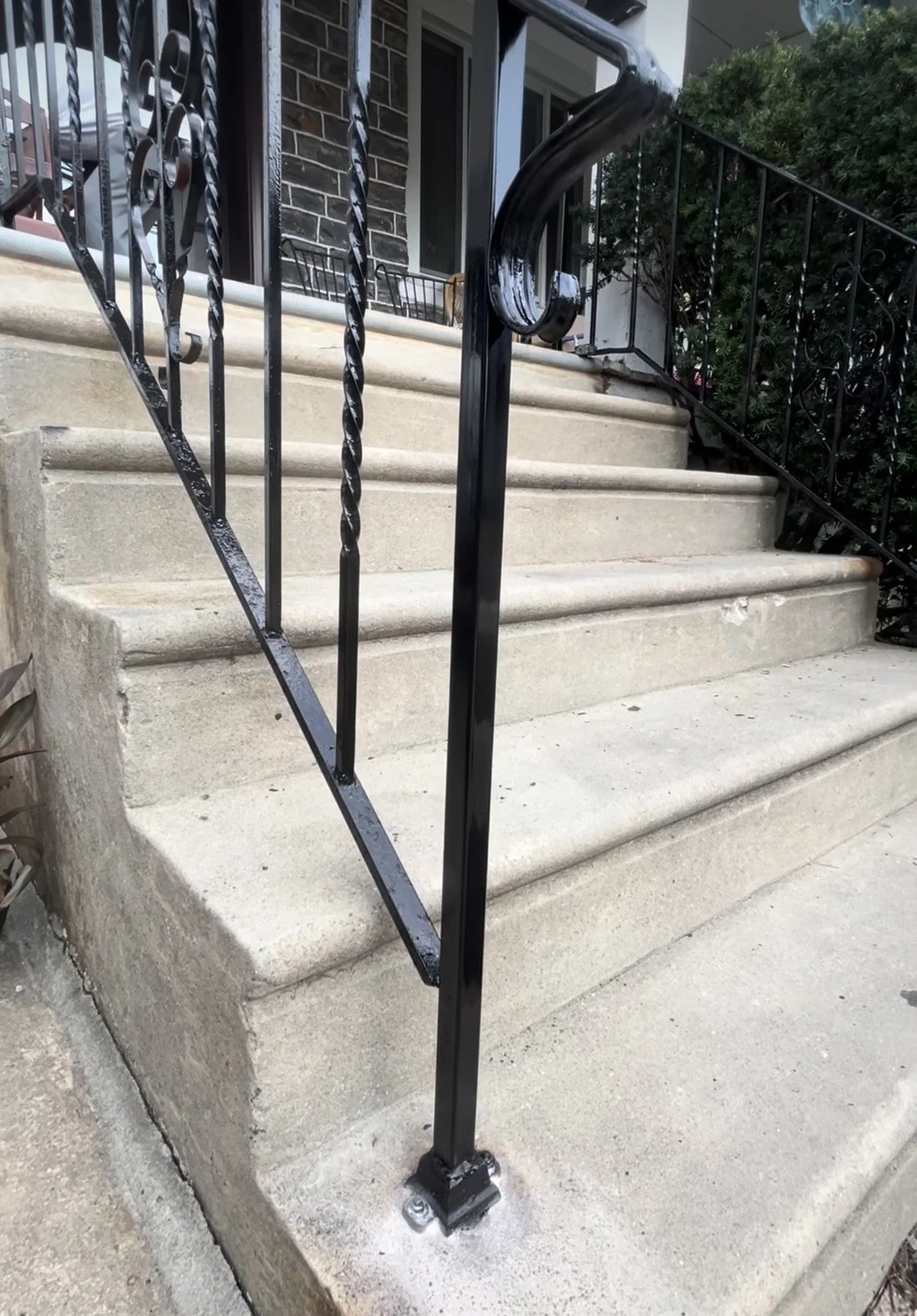 Close-up view of concrete stairs with a black metal handrail and a decorative balustrade, leading up to a house entrance with a window and green bushes nearby.