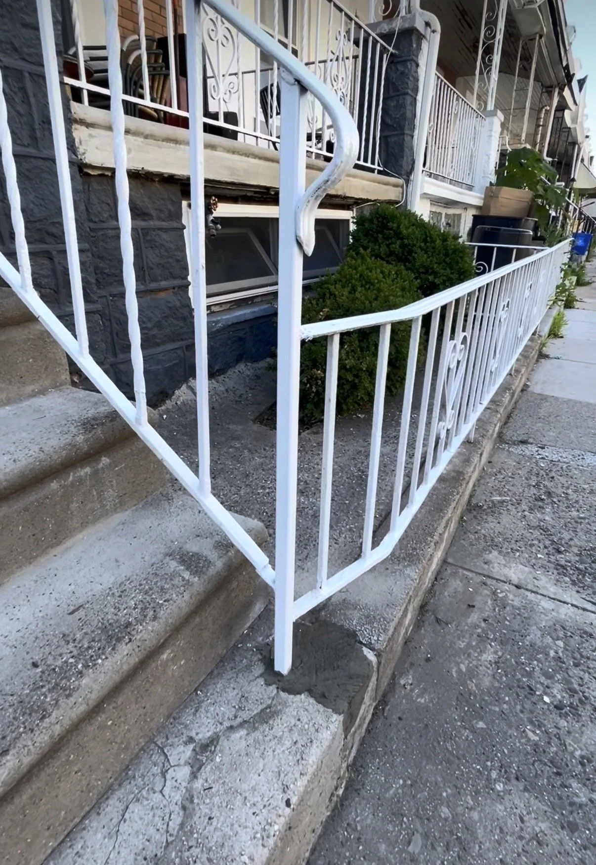 Close-up of a small front porch with a white metal railing surrounding concrete steps, with bushes and potted plants nearby, and part of a house visible in the background.