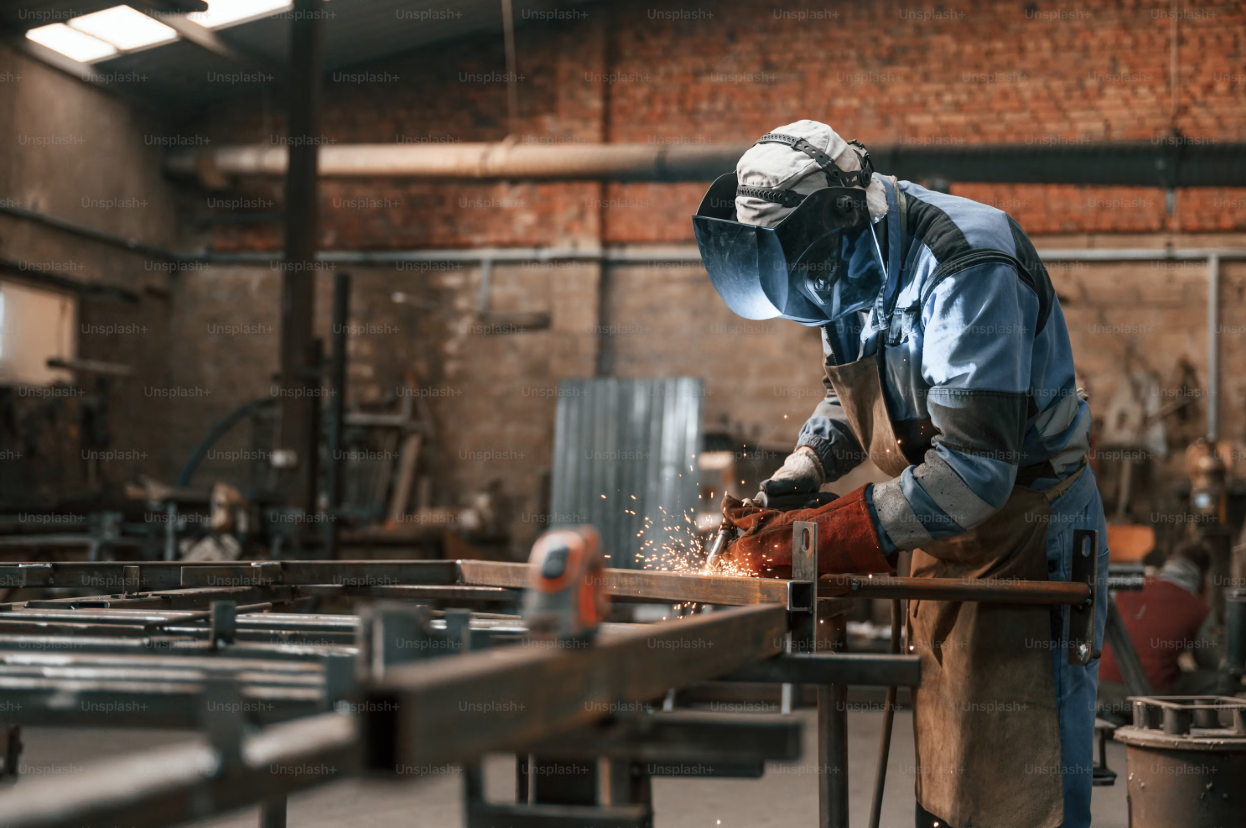 A person welding metal in a workshop with brick walls, wearing protective gear, including a mask, gloves, and apron.