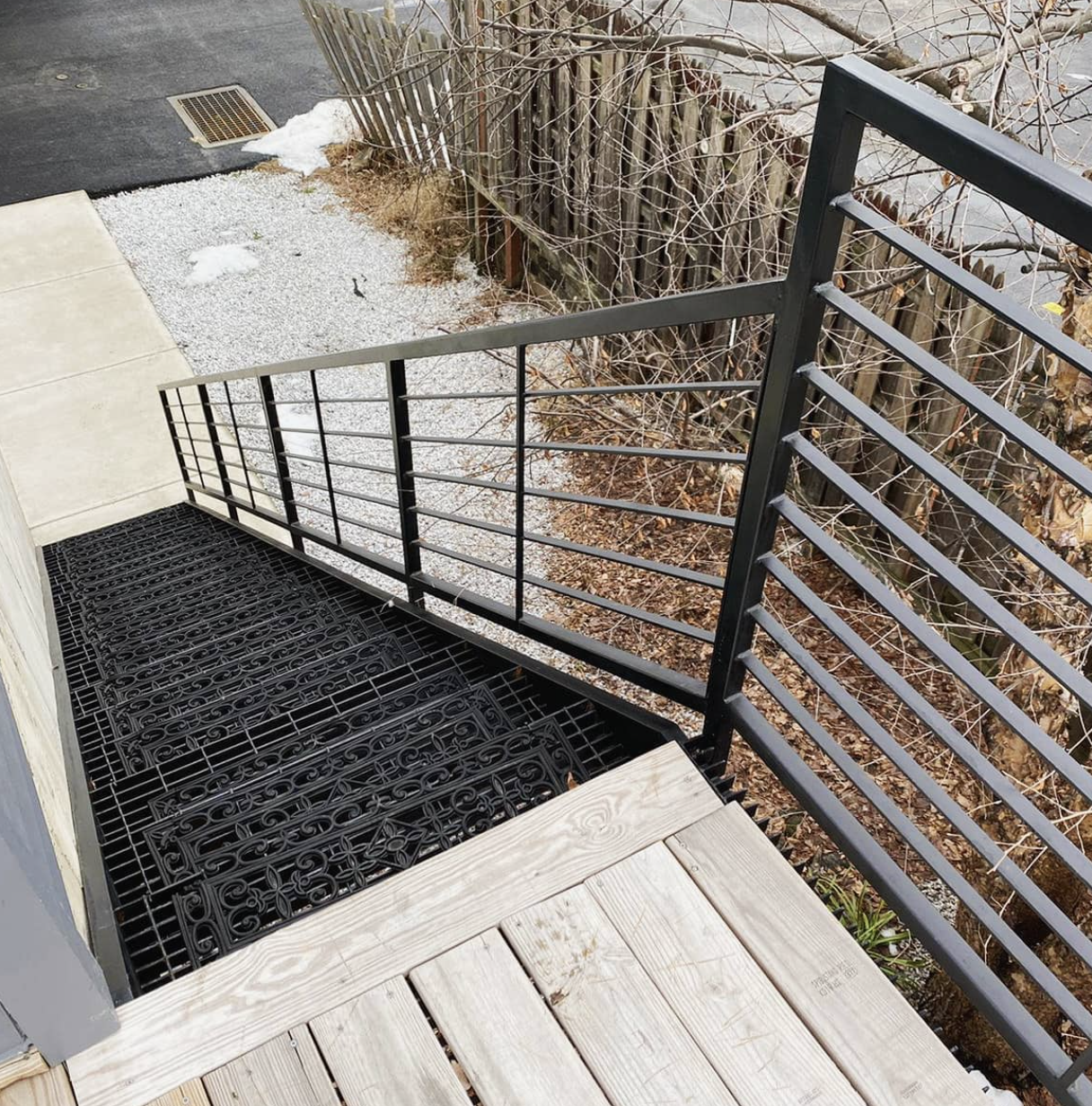 View of a small outdoor balcony with wooden floor, black metal railing, and decorative black mat or grate. Outside the railing, there are leafless bushes, a gravel area, and a concrete and asphalt surface with patches of snow.