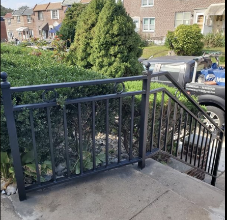 View from a porch showing a black metal railing, a sidewalk, a bush, and parked cars in a residential neighborhood.
