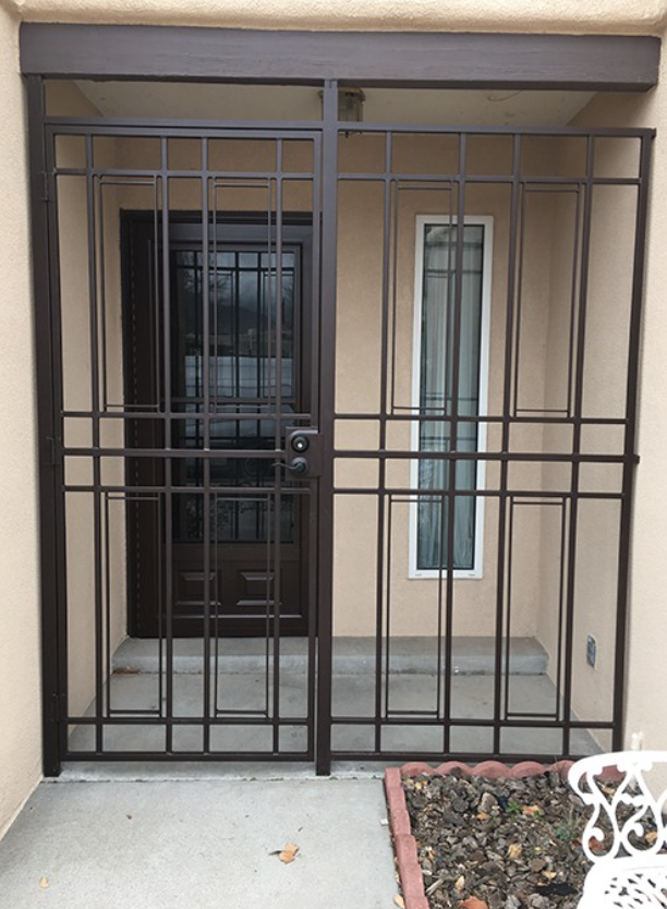 Front entrance with a metal security gate in front of a door and window, beige exterior wall, concrete floor, small garden bed with soil and a decorative white metal chair nearby.