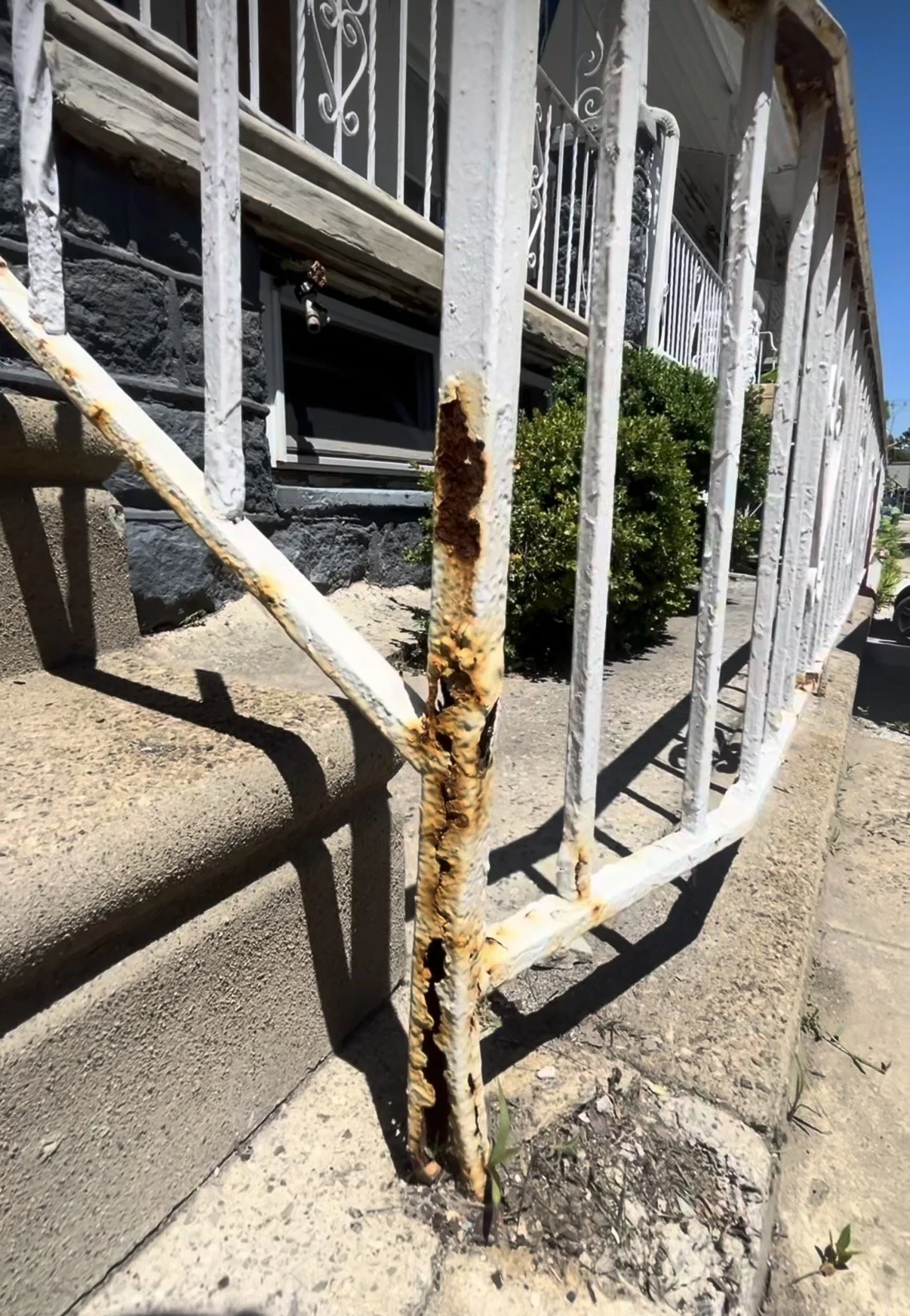 Rusty white metal fence in front of a house with black stone foundation, a small green bush, and a small window. The ground is dry and dirt-covered.