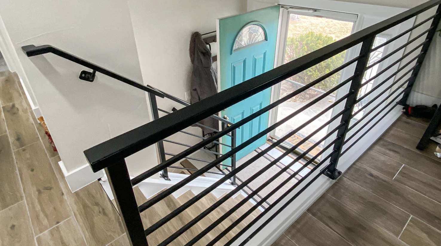 View from upstairs looking down at a black metal railing, front door painted blue, with some outdoor greenery visible through the open door.
