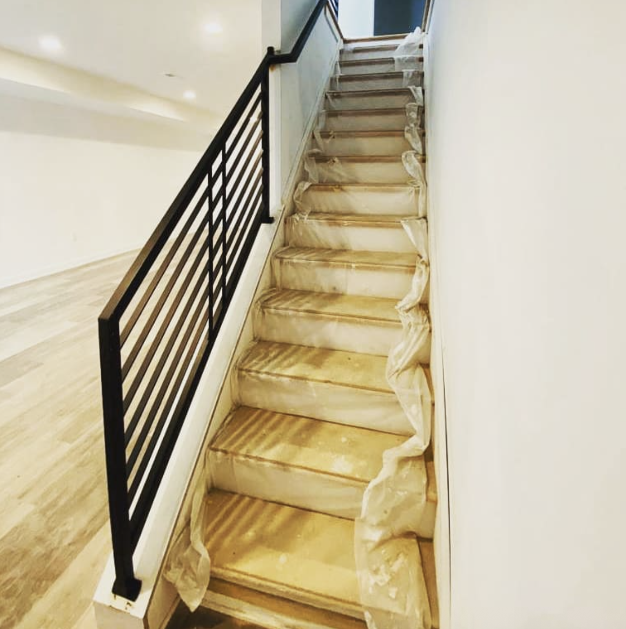 Indoor staircase under renovation with protective paper on the steps, black metal railing on one side, with a view of the upper level and window at the top.