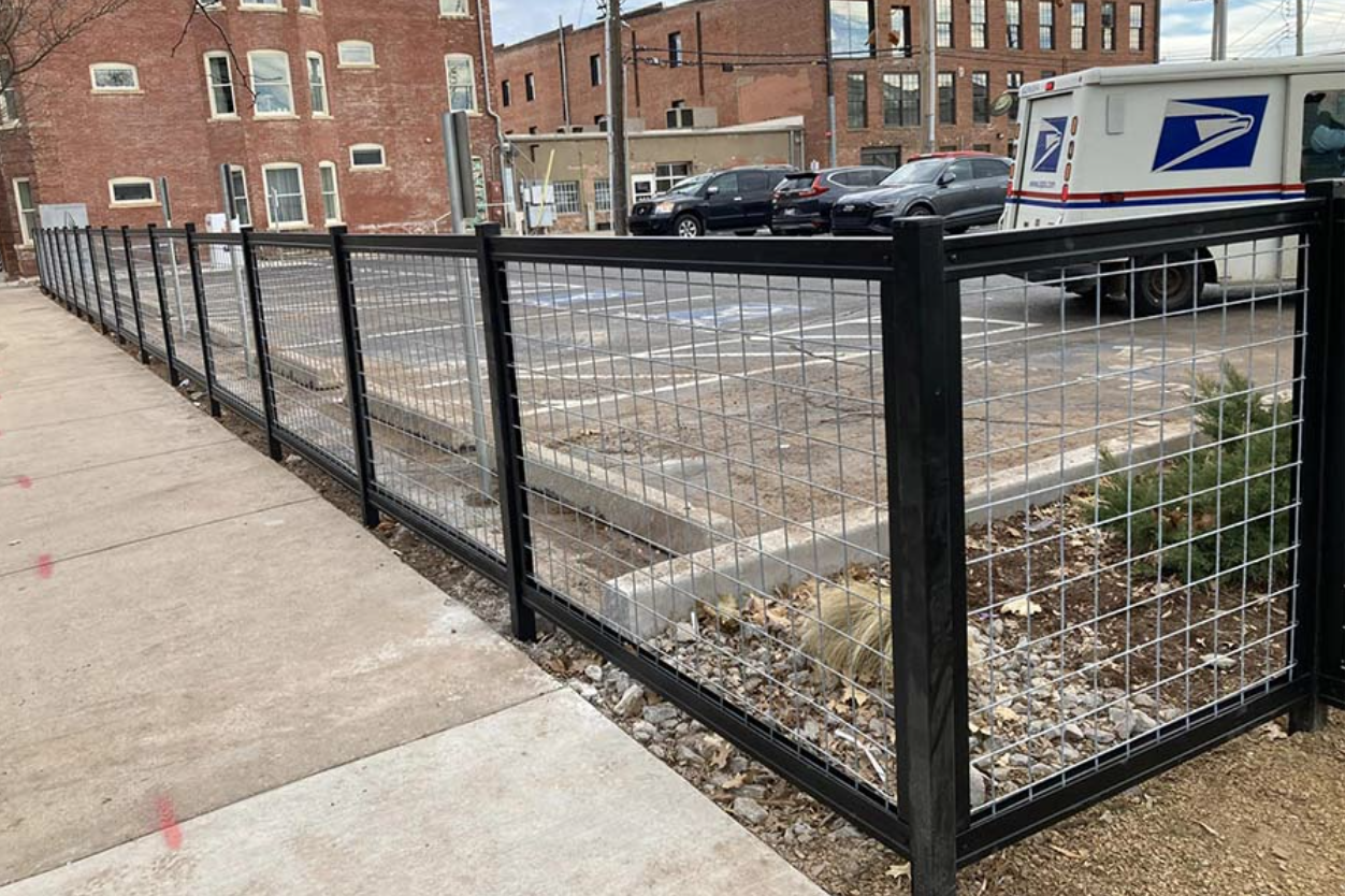 City sidewalk with a black metal fence, parking lot with cars, USPS mail truck, and brick buildings in the background.
