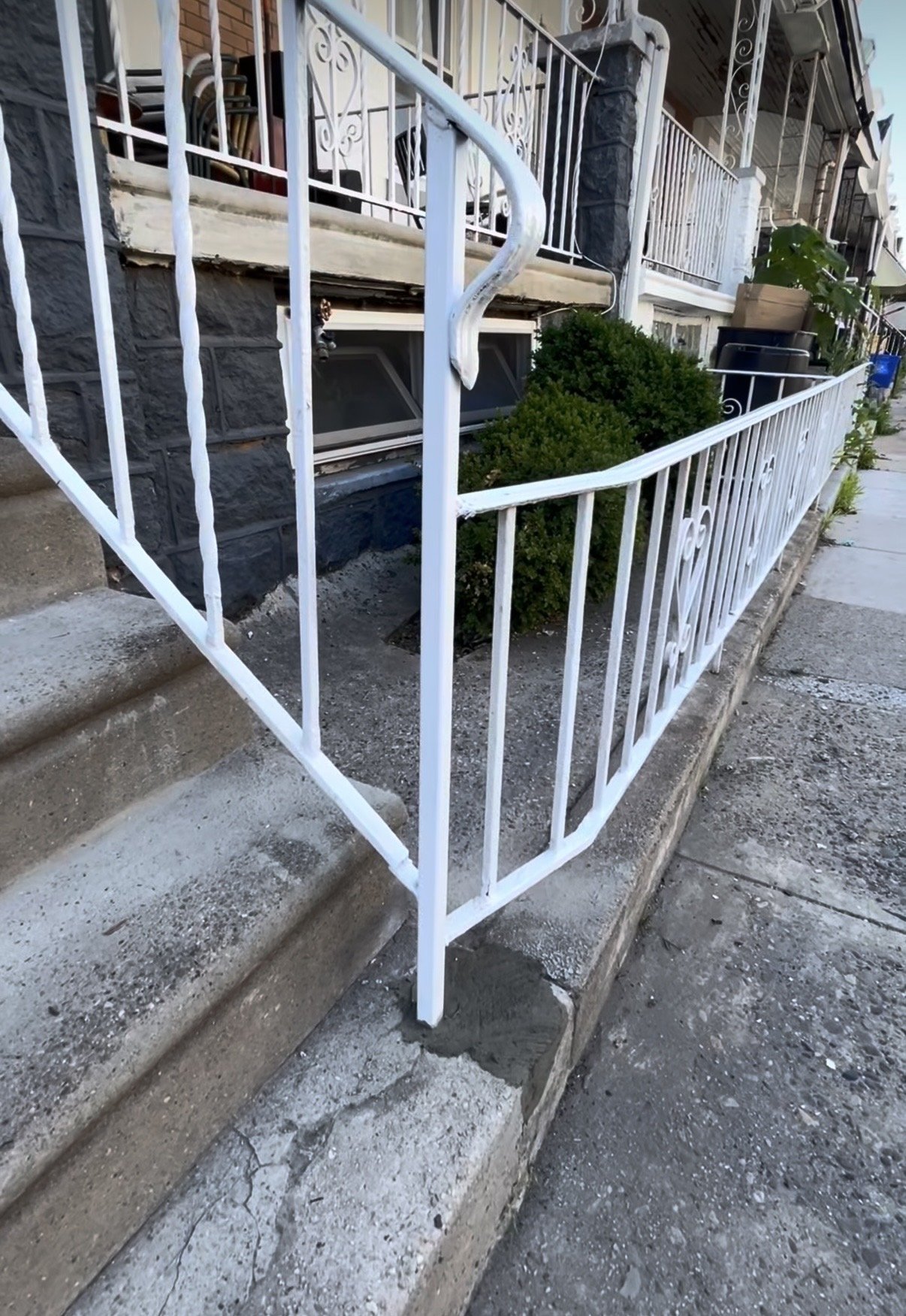 A front yard with concrete steps leading to a porch, enclosed by a white metal fence with decorative details. Small bushes are planted along the base of the porch, and there are potted plants and outdoor furniture visible on the porch.