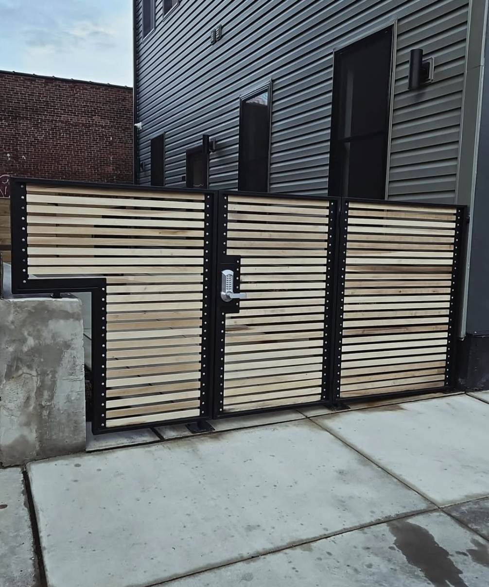 A wooden and metal gate with a keypad lock in front of a gray building with black window frames.