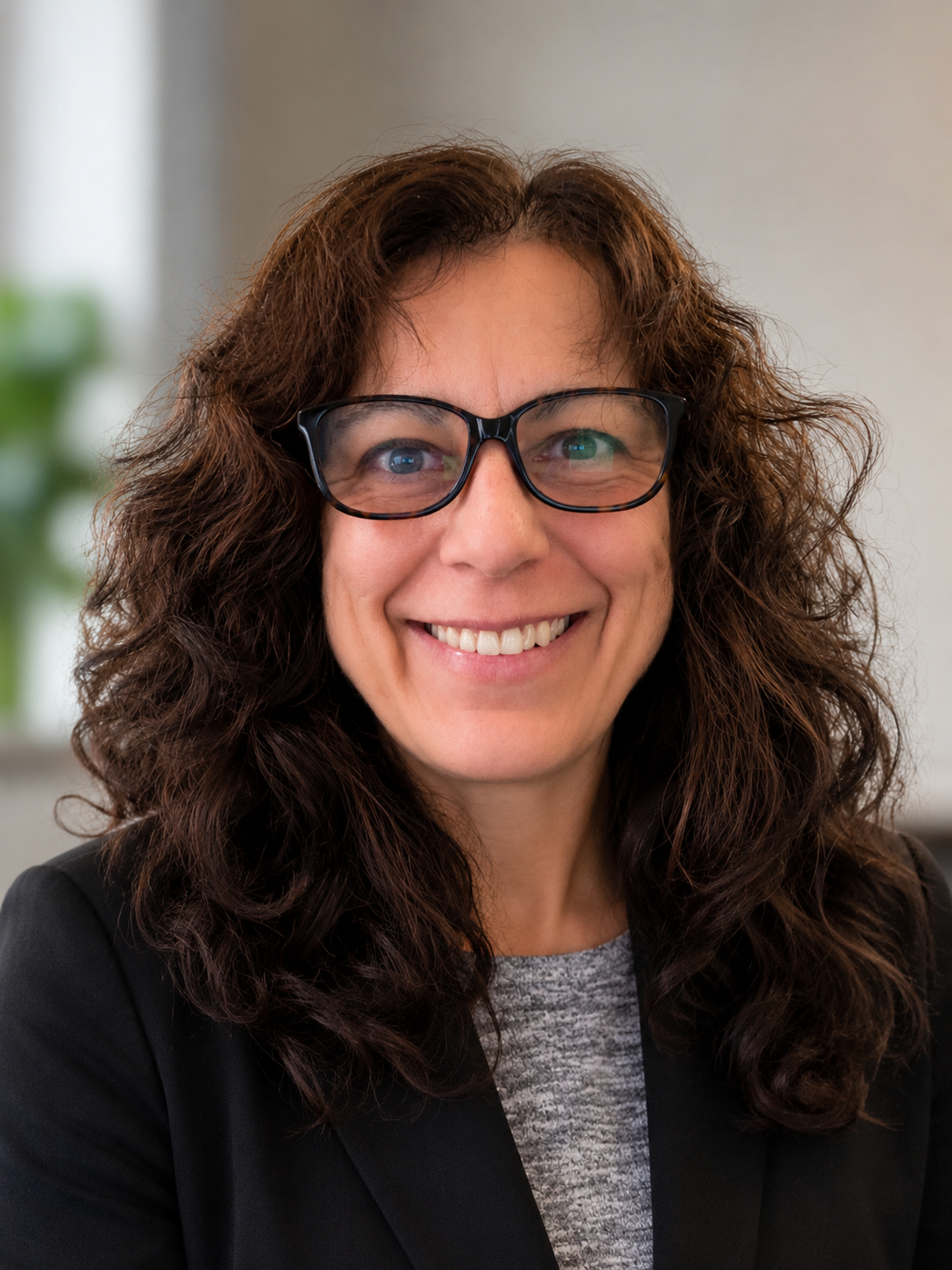 A woman with curly brown hair, wearing glasses, smiling, dressed in a black blazer and a gray top, with a blurred indoor background.