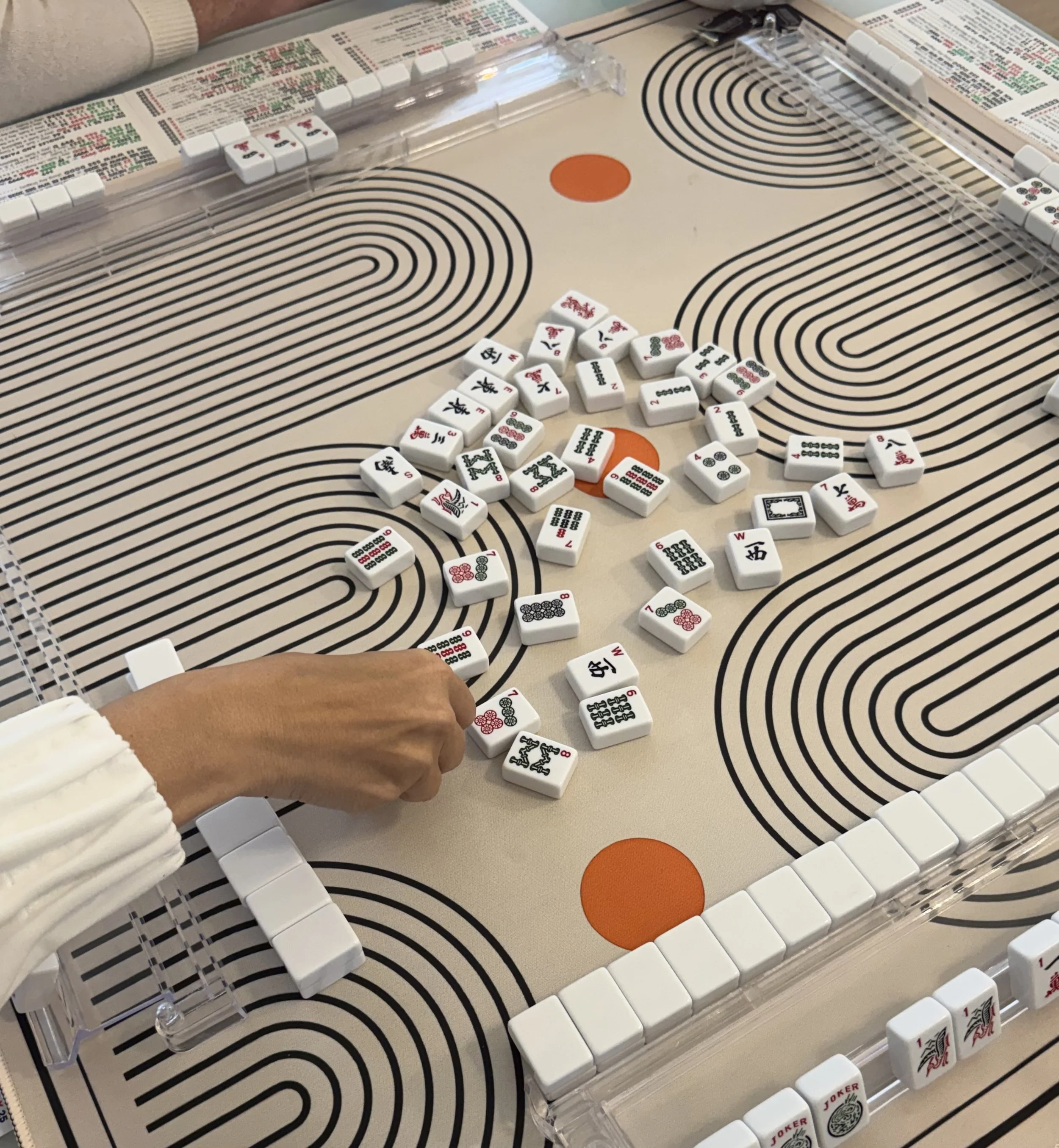 Close-up of a Mahjong game set with tiles arranged on a mat with concentric circle patterns, a person's hand reaching to pick a tile, and a transparent plastic tile rack.