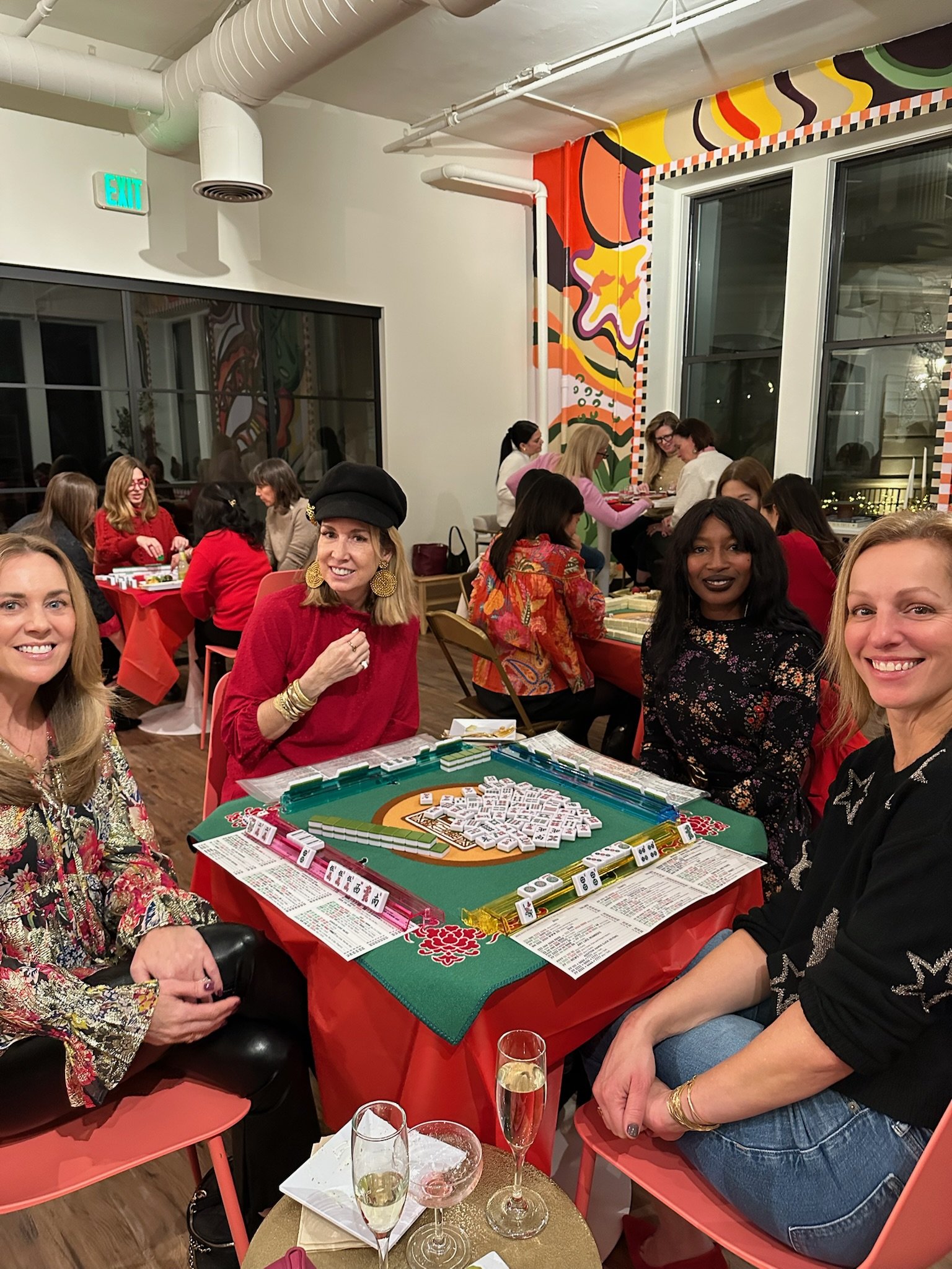 A group of women sitting around a table playing dominoes at a festive gathering. The table is decorated with a red tablecloth, and there are glasses of champagne and wine on a smaller side table. The room has colorful, abstract wall art, large windows, and a lively atmosphere.