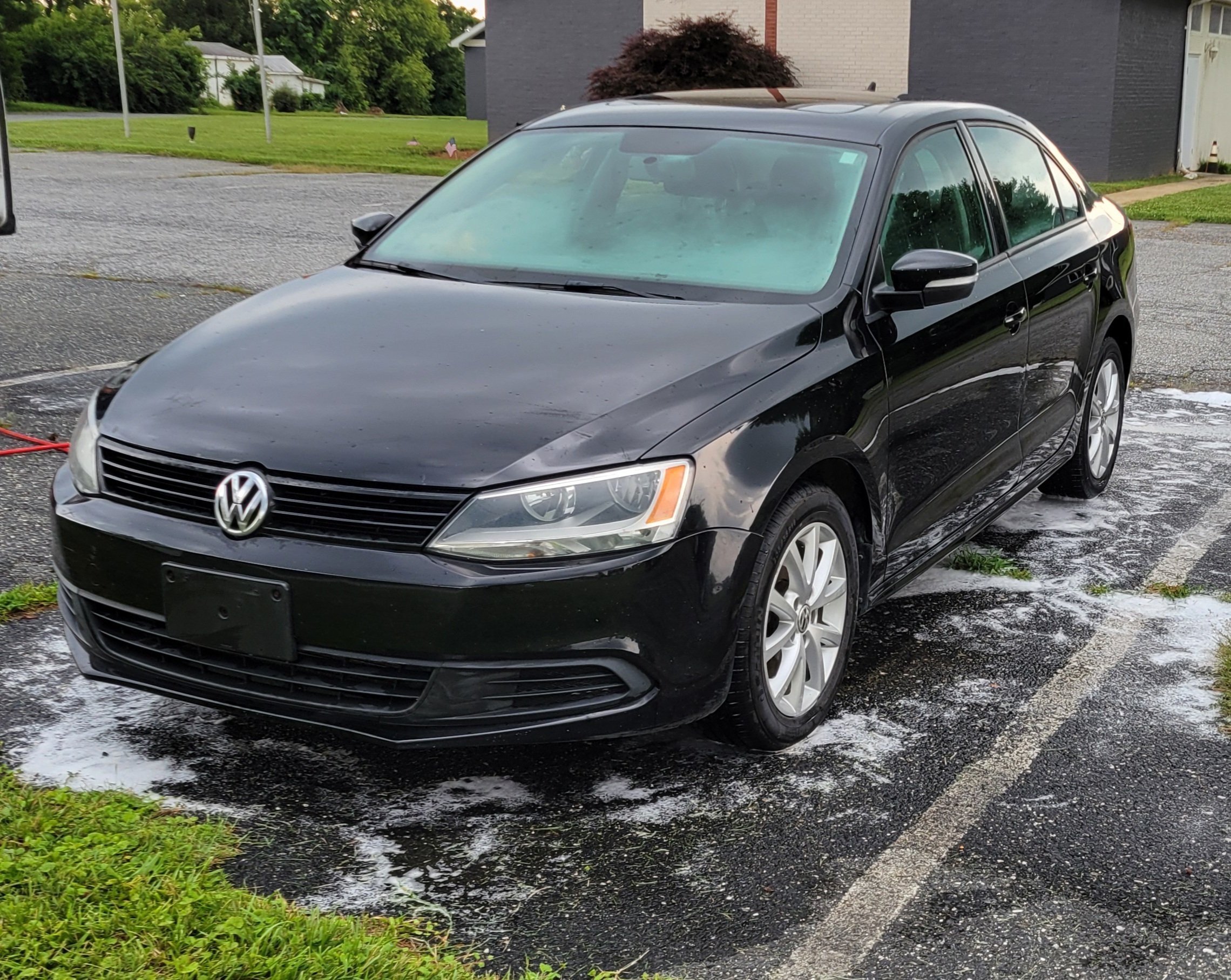 A black Volkswagen sedan parked on a wet driveway with soap and water nearby, in a residential area with green grass and trees.