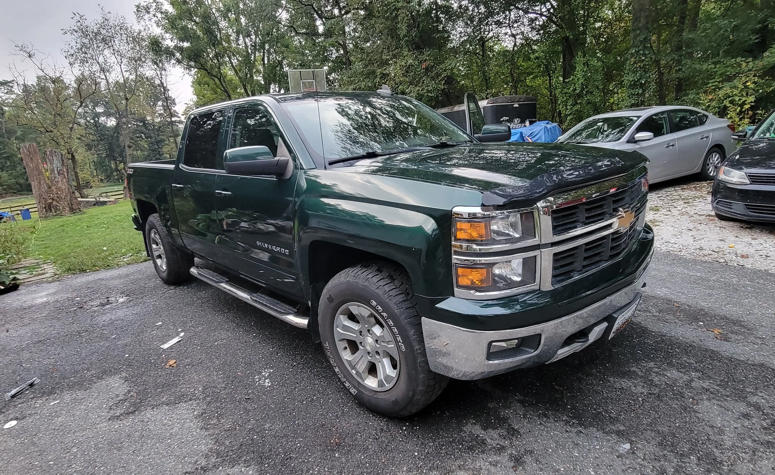 Rear view of a dark green Chevrolet Silverado pickup truck parked in a driveway with other cars around and trees in the background.