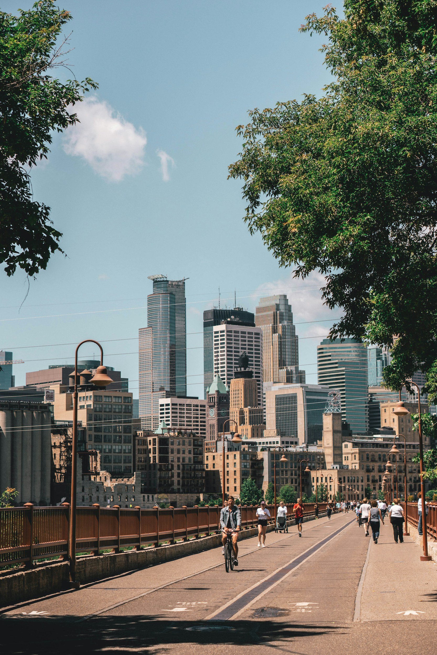 Cityscape with tall modern buildings, a bike lane with a cyclist, and pedestrians walking on a bridge with orange railings, trees on either side, under a clear blue sky with a few clouds.