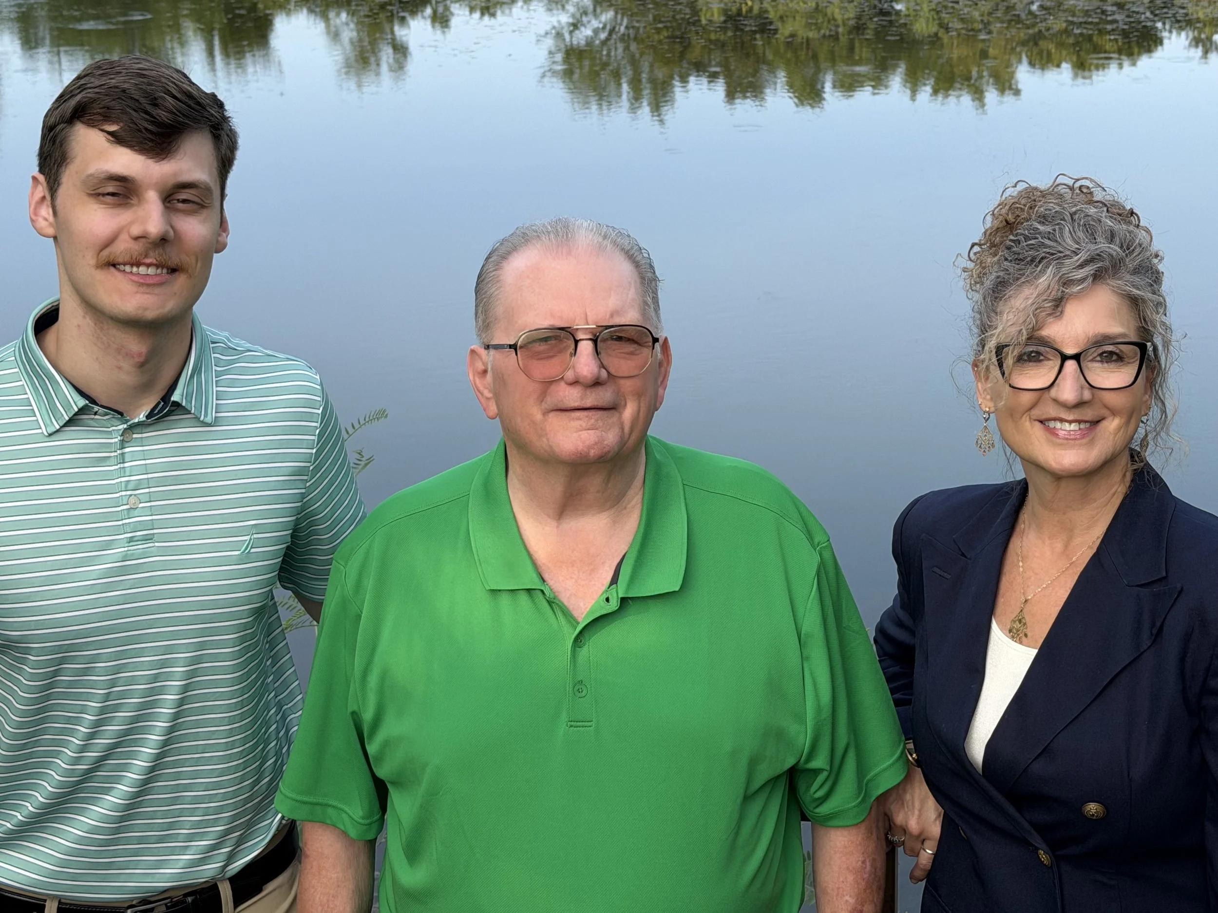 Three people standing outdoors near a body of water, with trees reflected in the water, smiling and looking at the camera.