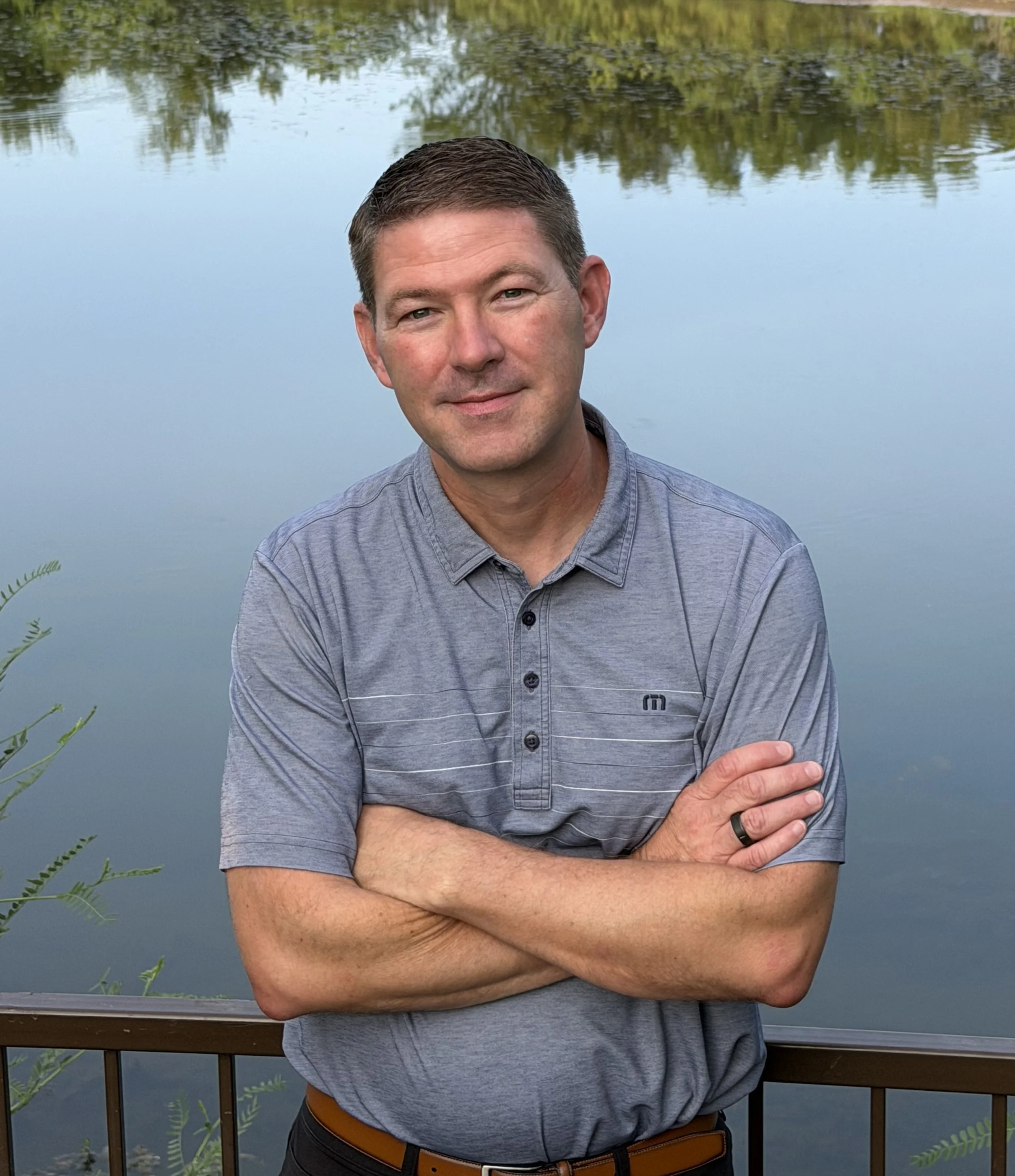A man with short brown hair and a slight smile, wearing a gray button-up shirt with a logo, standing with arms crossed near a body of water with reflections of trees, on a wooden deck with some greenery in the foreground.