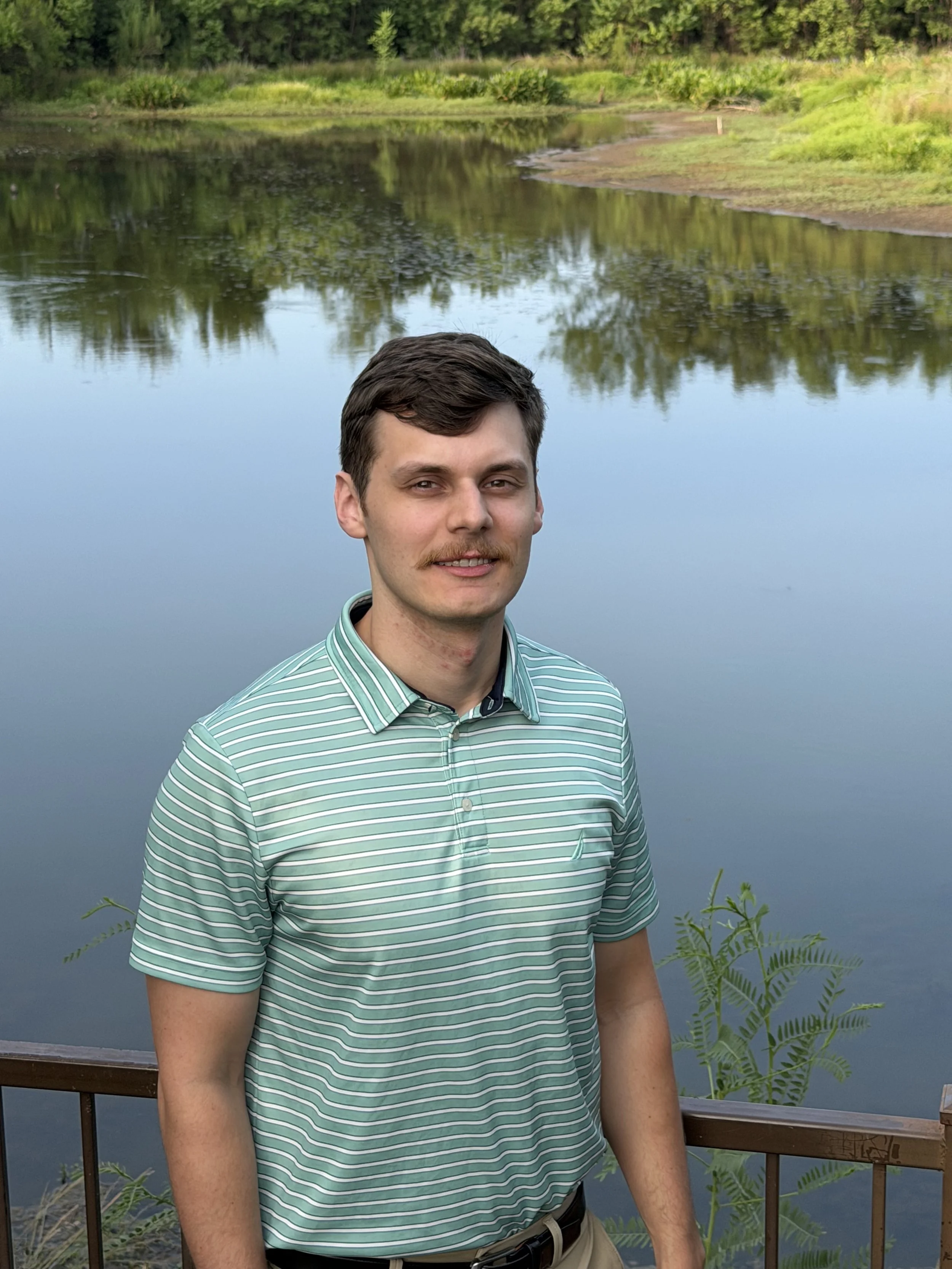 A young man with dark hair and a mustache, wearing a green and white striped polo shirt, standing outdoors near a body of water with greenery and trees in the background.