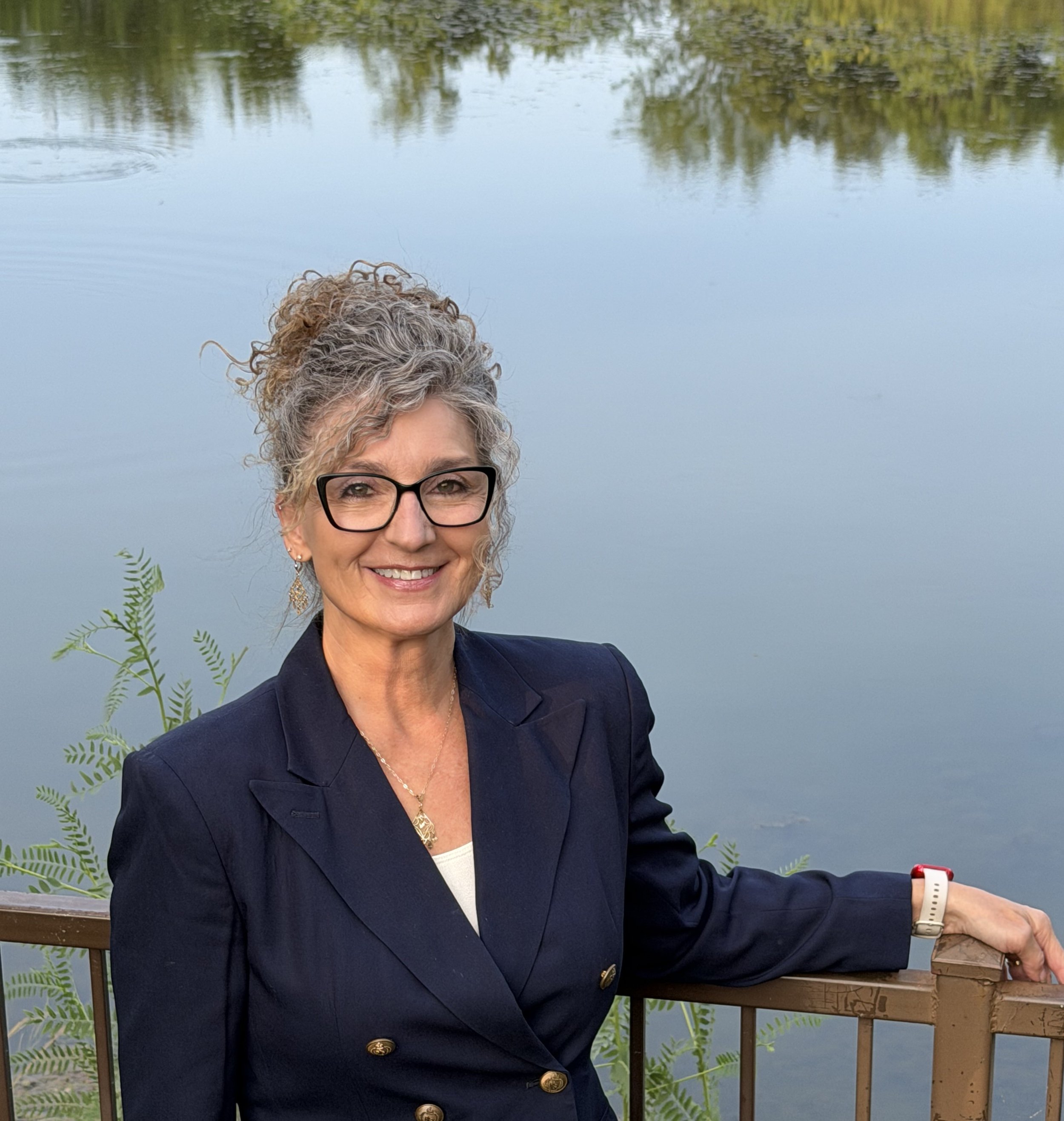 A woman with curly gray hair and glasses, smiling, wearing a navy blue blazer with gold buttons, standing outdoors by a lake with a wooden railing and green foliage in the background.