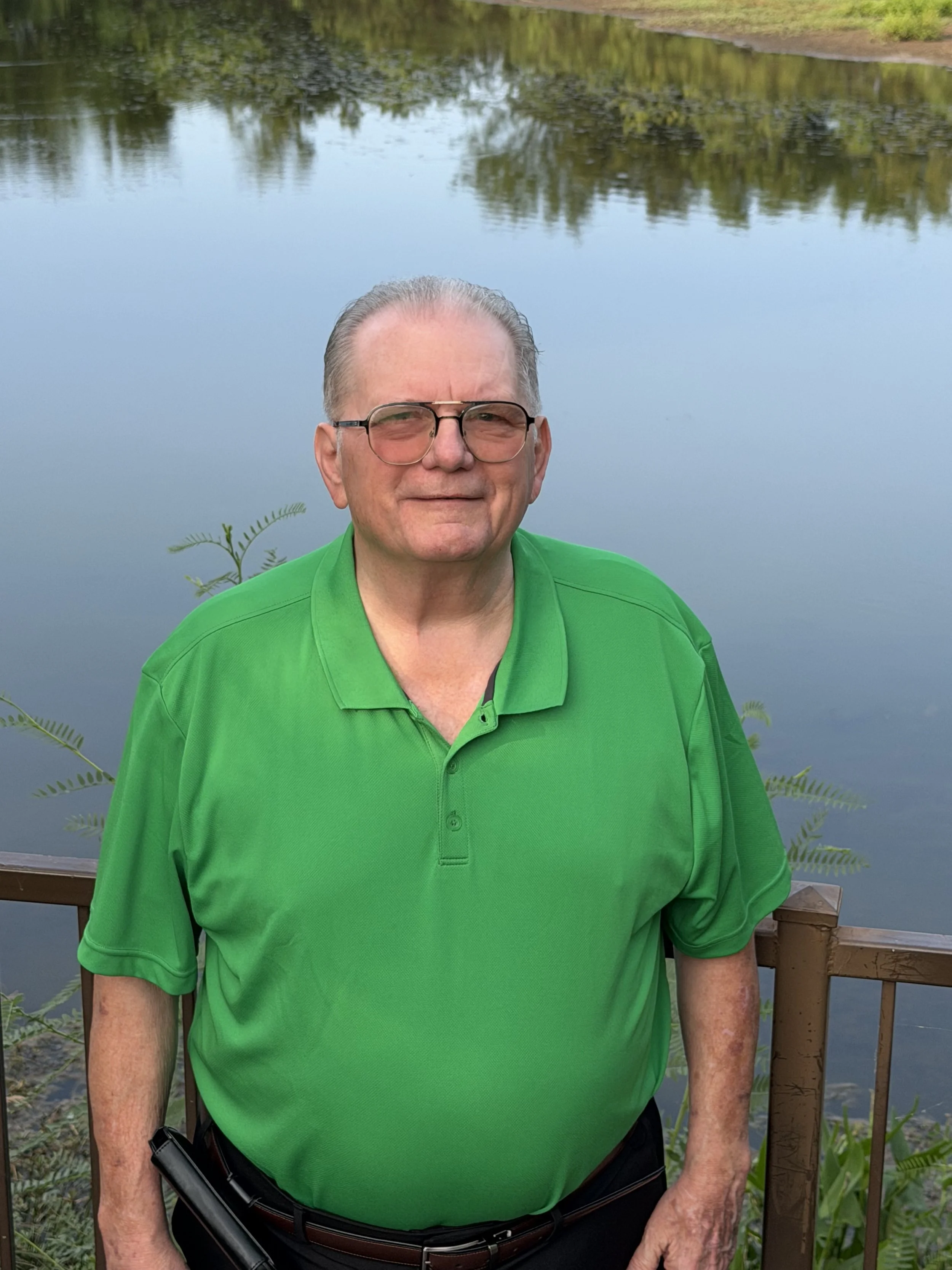 An elderly man with glasses wearing a bright green polo shirt standing outdoors near a body of water and surrounded by greenery.
