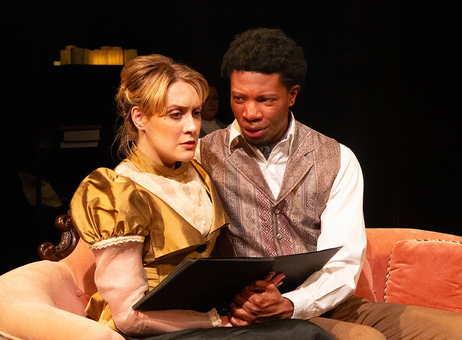 Production photo of Hedda Gabler. Two actors sitting and looking at a photo album with serious expressions. The woman has blonde hair in an old-fashioned gold dress with puffed sleeves, and the man has black curly hair in a vest and white shirt.