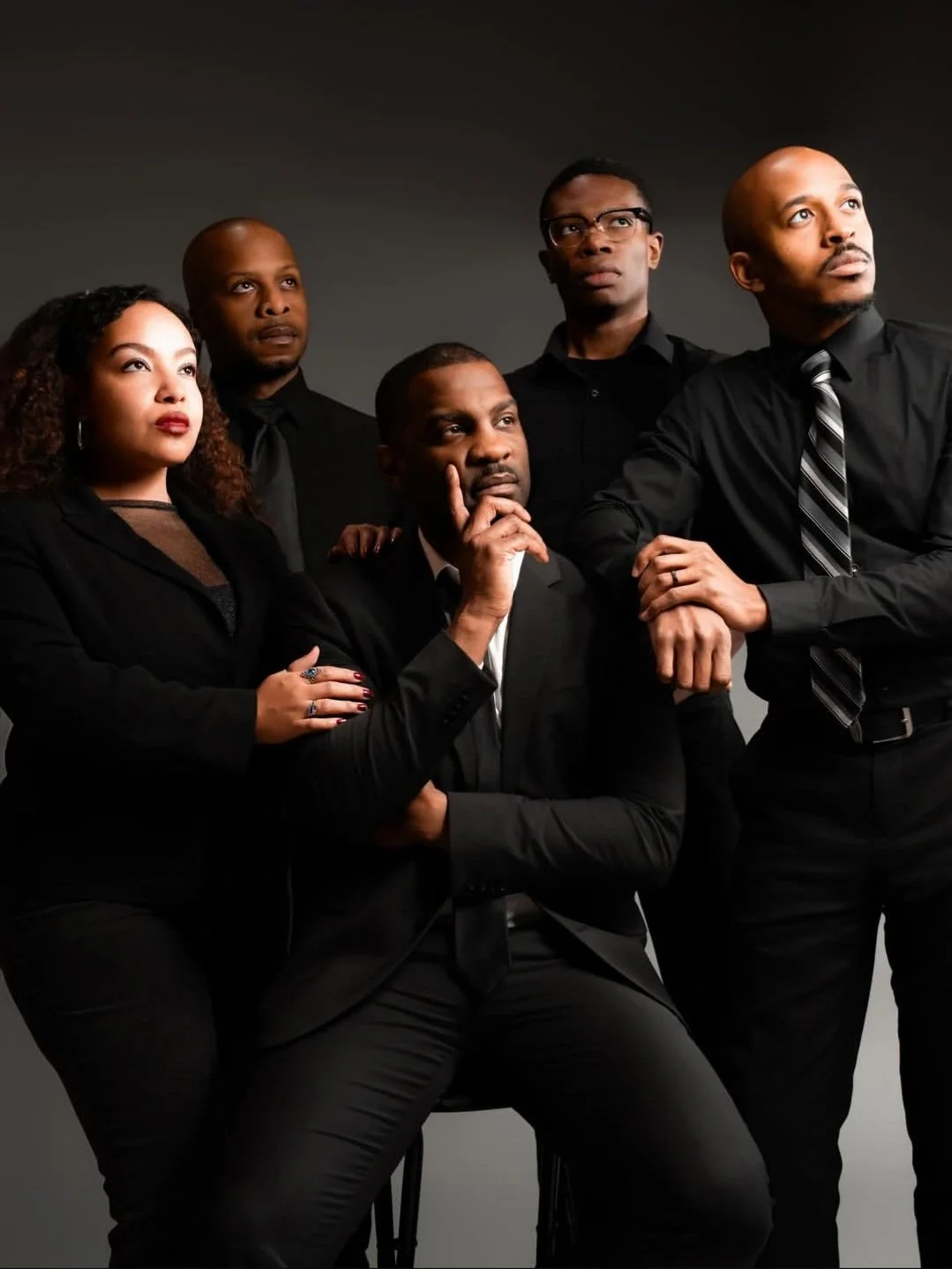 Cast photo of Stokely and Martin. A group of five Black people dressed in formal black attire, with serious expressions, posing against a dark background.
