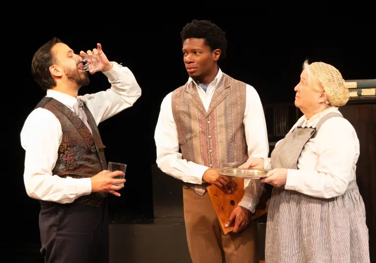 Production photo of Hedda Gabler. Three actors on stage in period costumes, with one drinking from a glass, another holding a plate and a musical instrument, and the third observing, against a dark background.