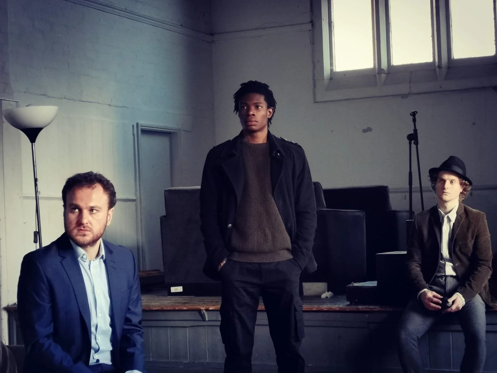 Three men sitting and standing in an industrial-style room with high windows and a microphone stand. One man is in a blue suit, another man is standing in the middle wearing a black jacket, and the third man is sitting on the right dressed in a brown