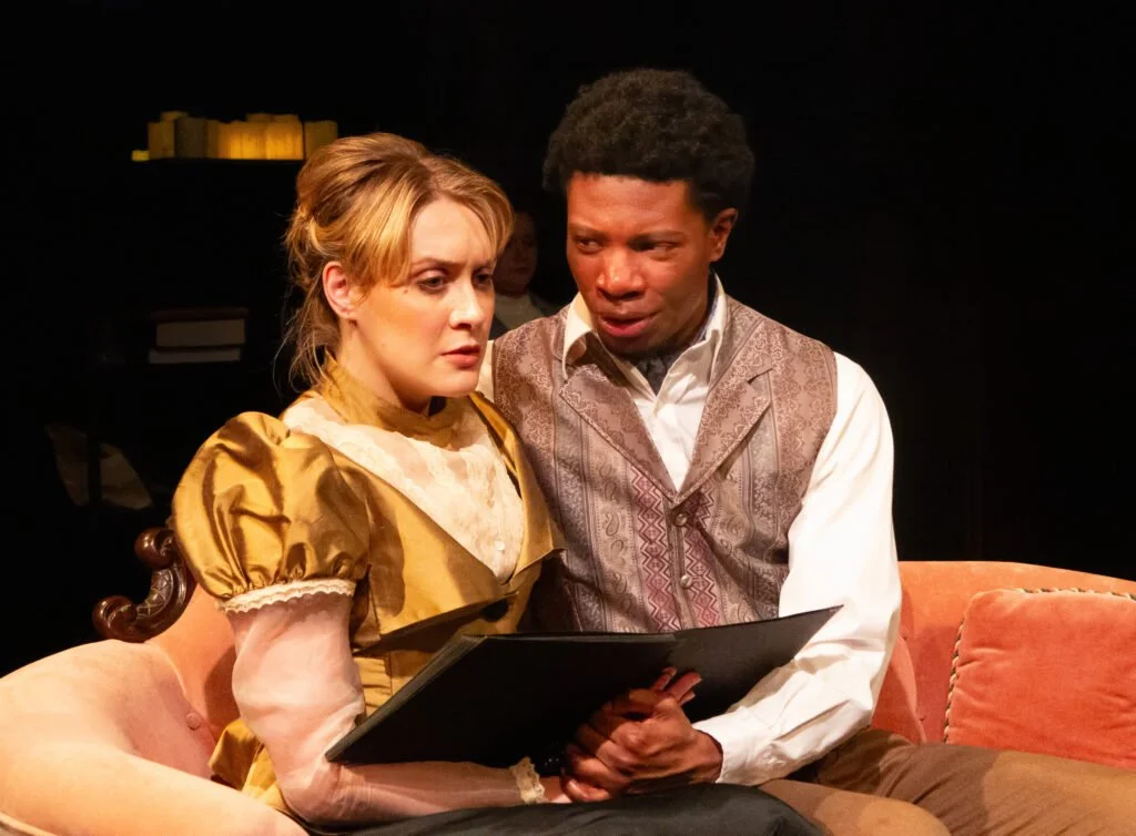Production photo of Hedda Gabler. Two actors on stage, a woman with blonde hair in a golden dress, and a man with short curly black hair in period costume, looking at a book or script together in a theater setting.