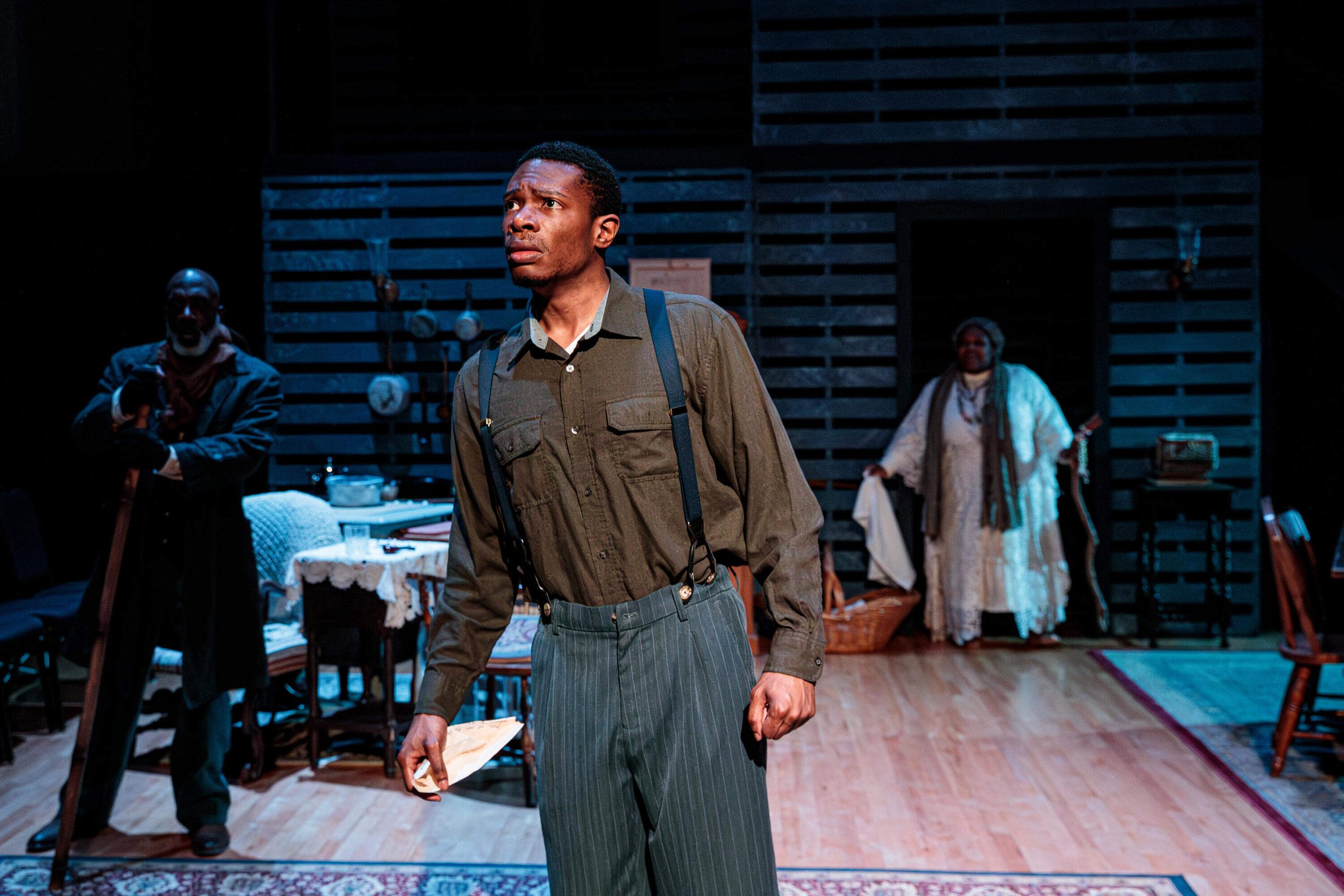 Production photo of Gem of the Ocean. Actors on stage performing in a play, with one man in the foreground wearing suspenders and holding a paper, and two others in the background behind a table with dishes, set in a room with wooden walls.