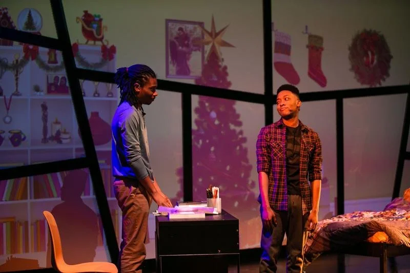 Production photo of Black Super Hero Magic Mama. Two boys stand in a room with images of a Christmas tree and stockings projected on the wall behind them. One boy is leaning on a desk, while the other looks at him with a thoughtful expression. 