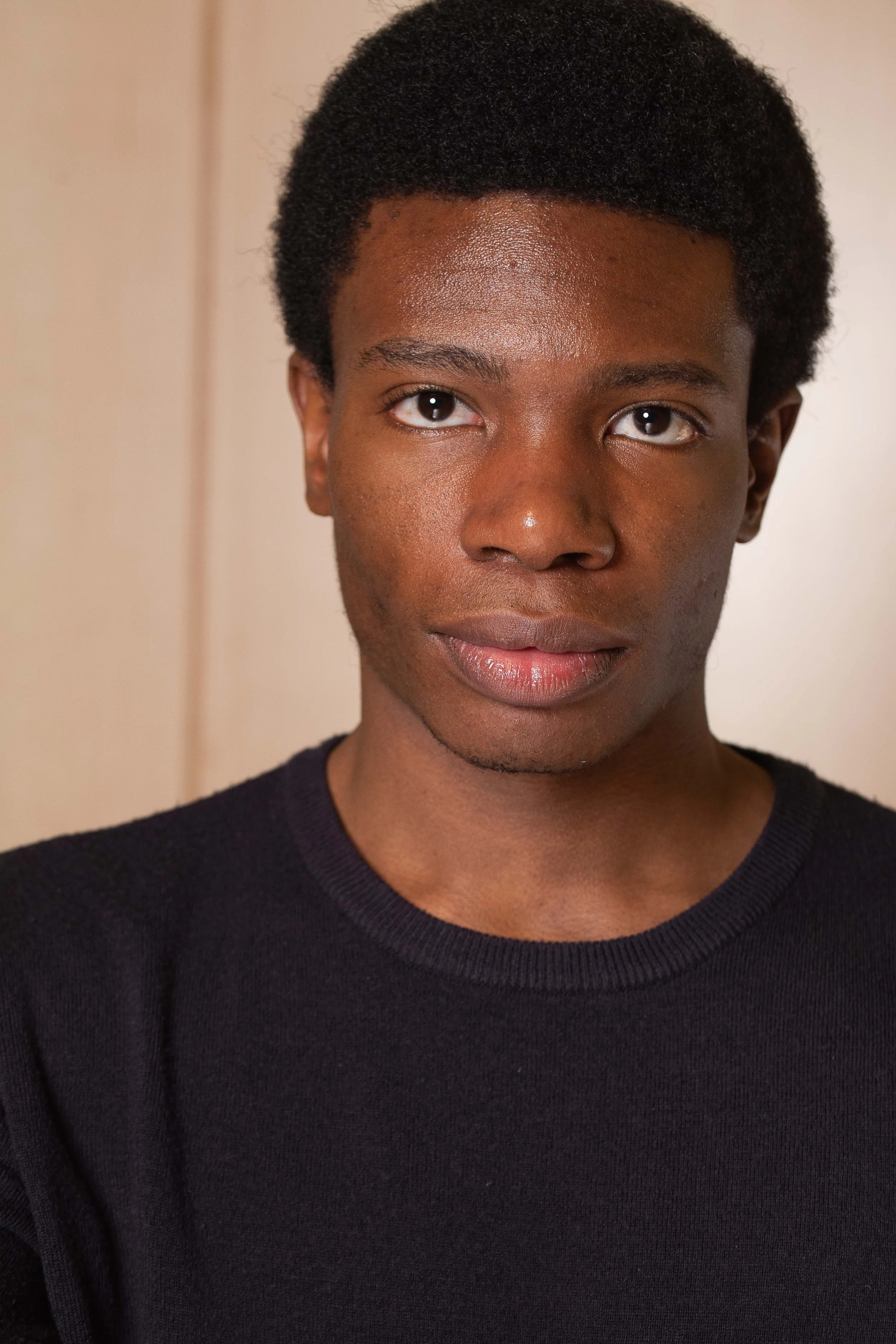 Close-up of a young person with dark skin, short curly hair, and wearing a black shirt, standing in front of a neutral background.