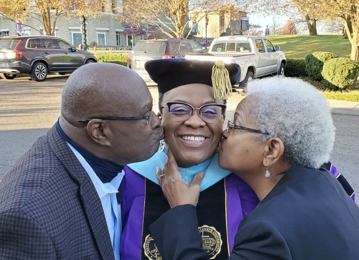 A young woman in a graduation cap and gown smiling as she is kissed on each cheek by an elderly man and woman, outside in a parking lot with cars and trees in the background.