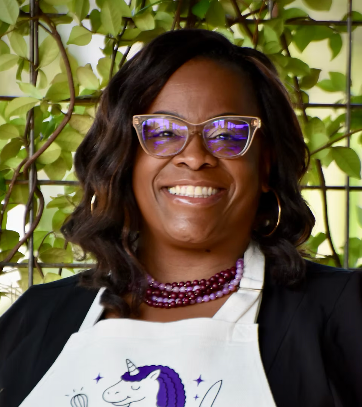 A woman smiling, wearing glasses with purple reflective lenses, a beaded purple necklace, and hoop earrings, standing in front of green leafy background.